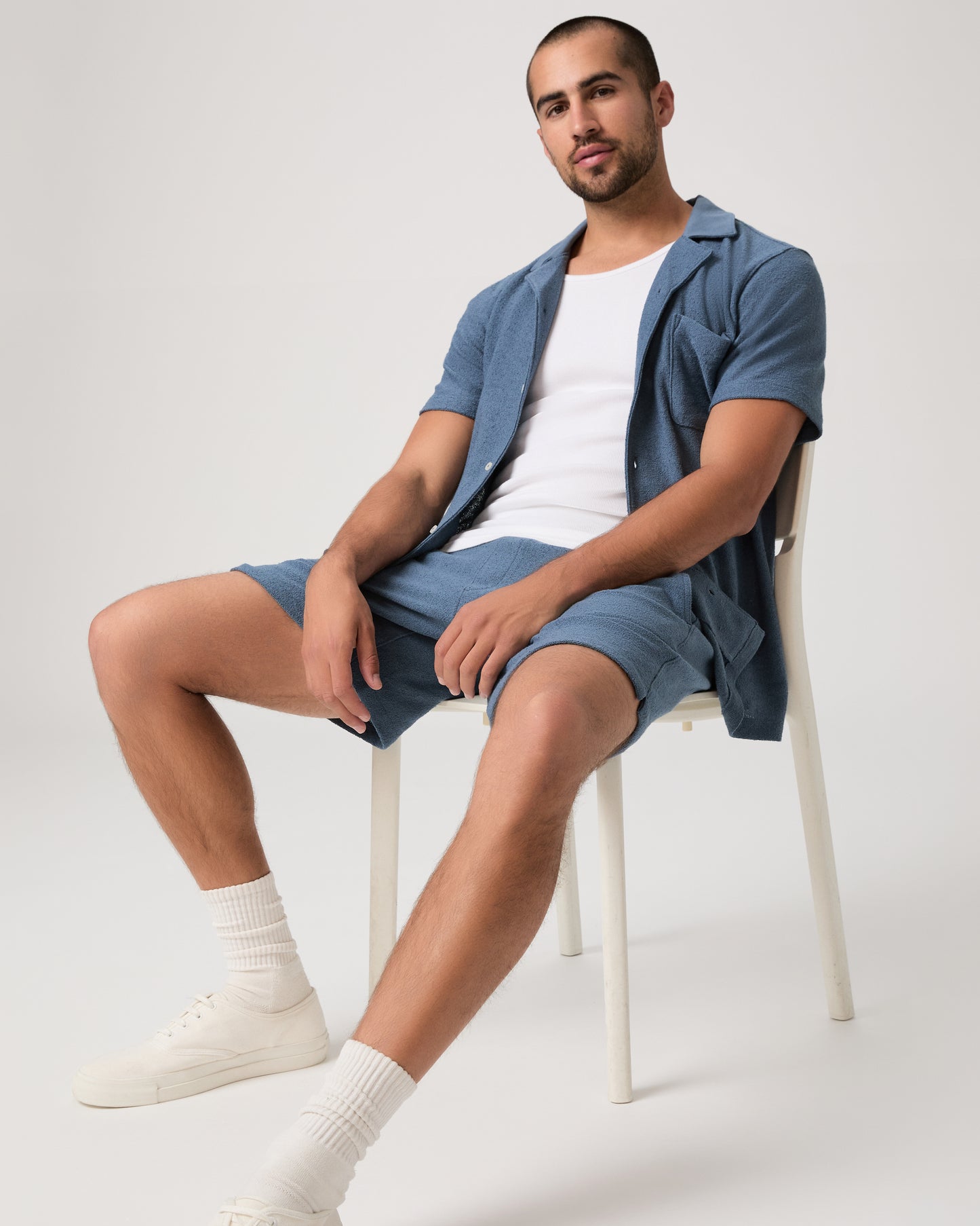 Man seating on a chair wearing a white under a blue shirt and shorts set with white shoes on a light gray background