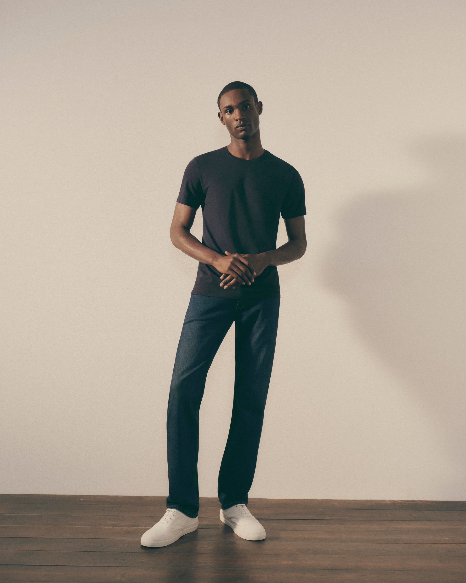 Man wearing a black t-shirt and dark pants standing against a plain wall.