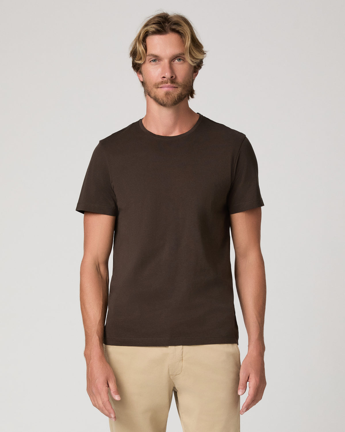 A man wearing a dark brown crew-neck t-shirt and beige pants, standing against a light gray studio background.