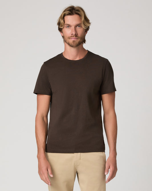 A man wearing a dark brown crew-neck t-shirt and beige pants, standing against a light gray studio background.