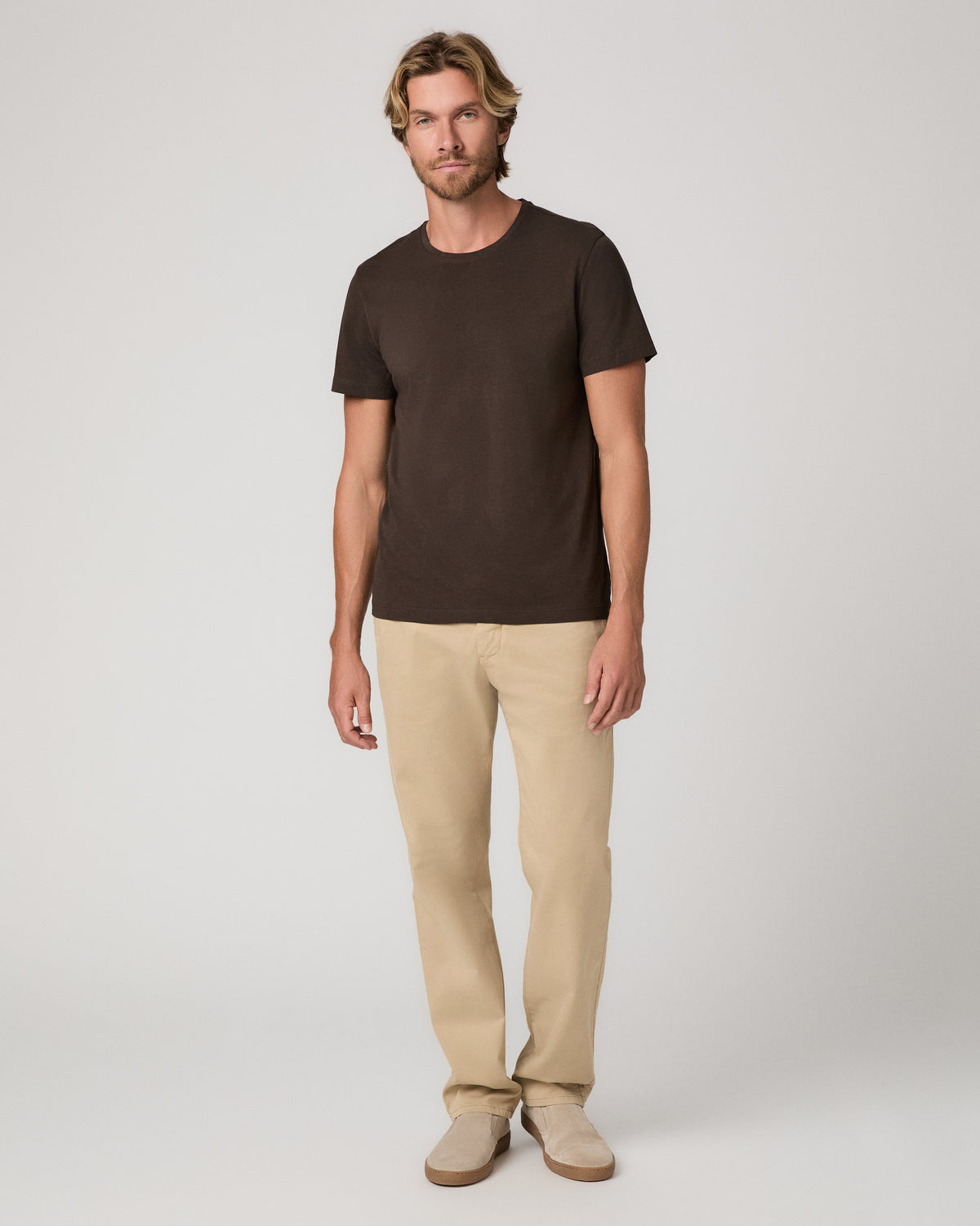 A man wearing a dark brown crew-neck t-shirt and beige pants, and beige shoes standing against a light gray studio background.