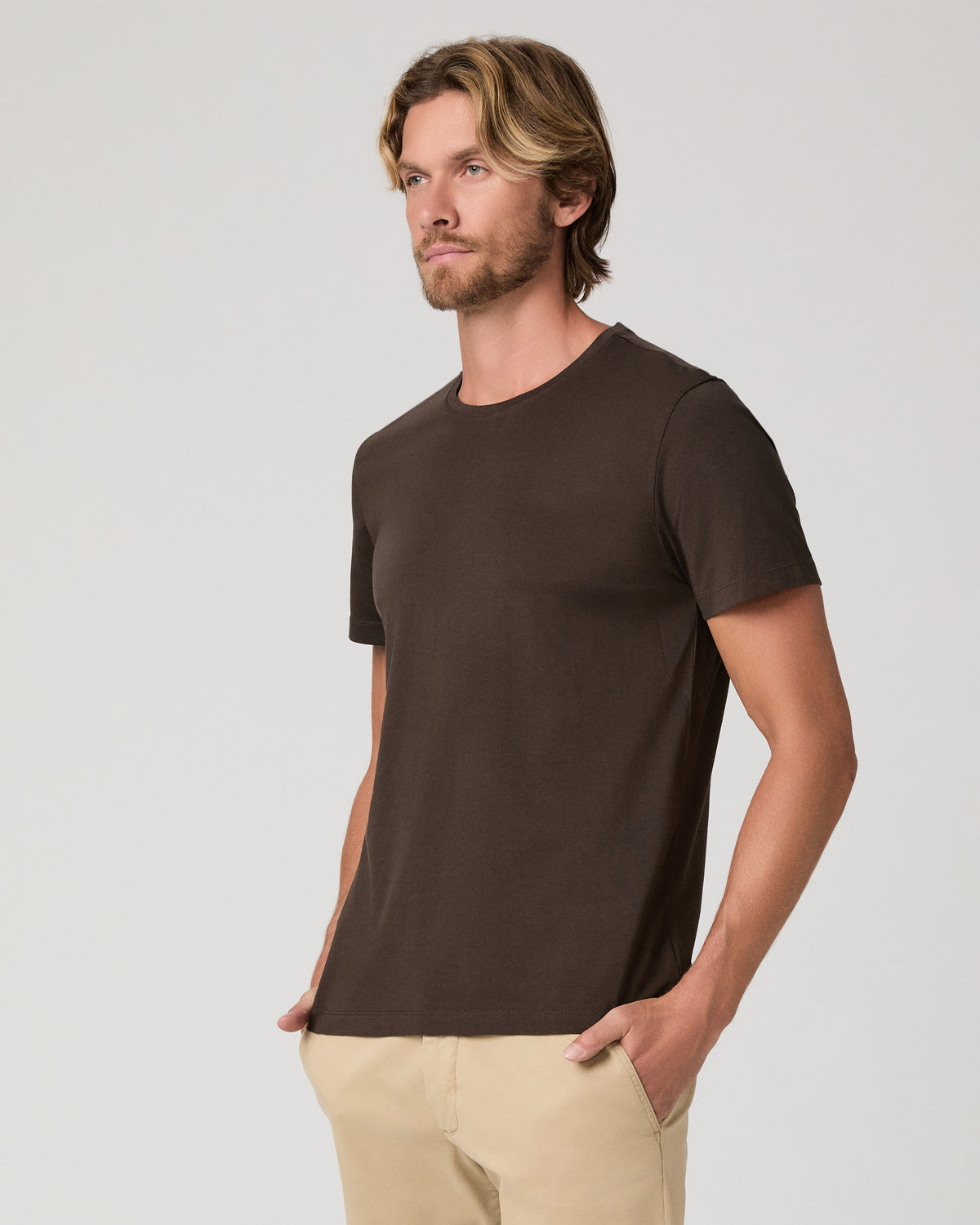 A man wearing a dark brown crew-neck t-shirt and beige pants, standing against a light gray studio background.
