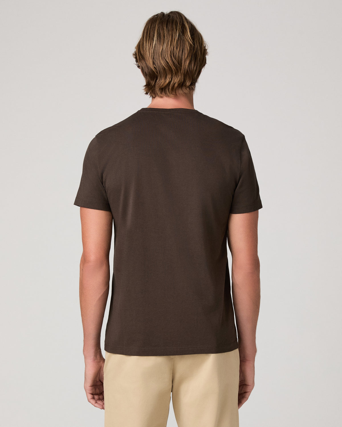 A man wearing a dark brown crew-neck t-shirt and beige pants, standing against a light gray studio background.