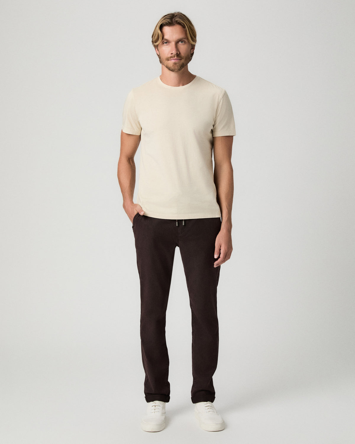 Full-length view of model with one hand in his pocket, wearing the Sorden Crew Neck Tee in Light Pale Antler with dark brown pants and white sneakers
