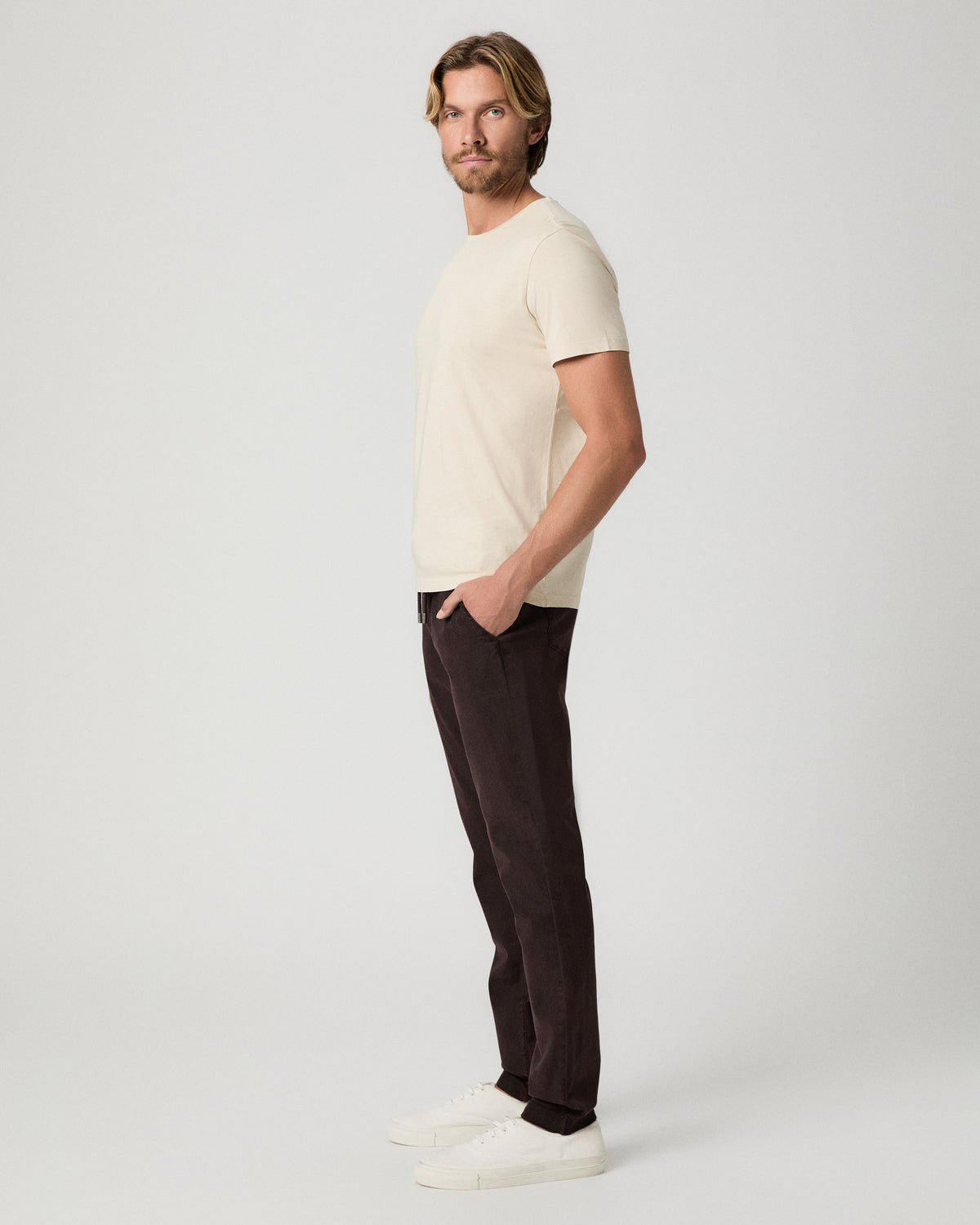 Side profile of model with one hand in his pocket, wearing the Sorden Crew Neck Tee in Light Pale Antler with dark brown pants and white sneakers