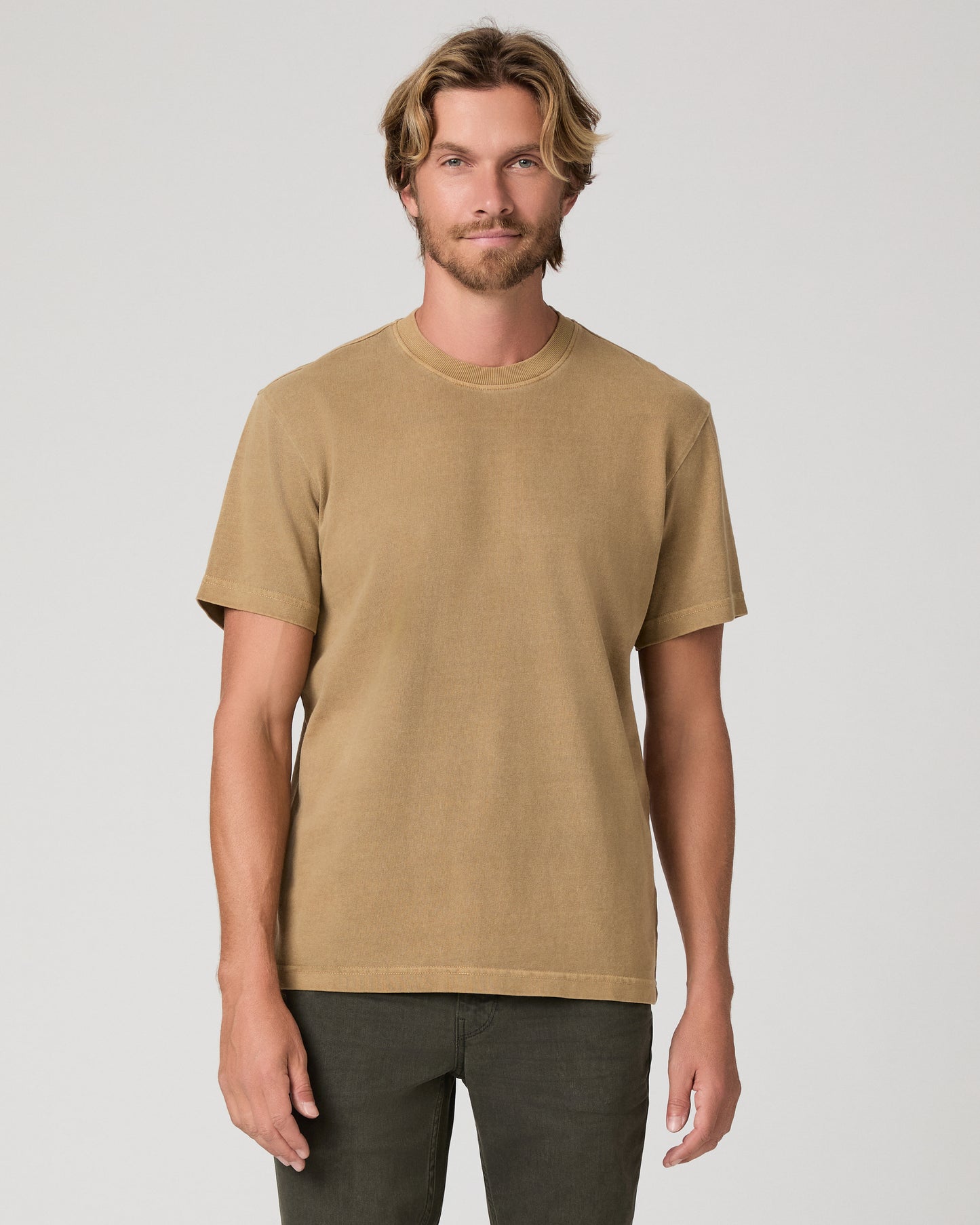 A man modeling a brown tee and dark green trousers against a light gray studio background.