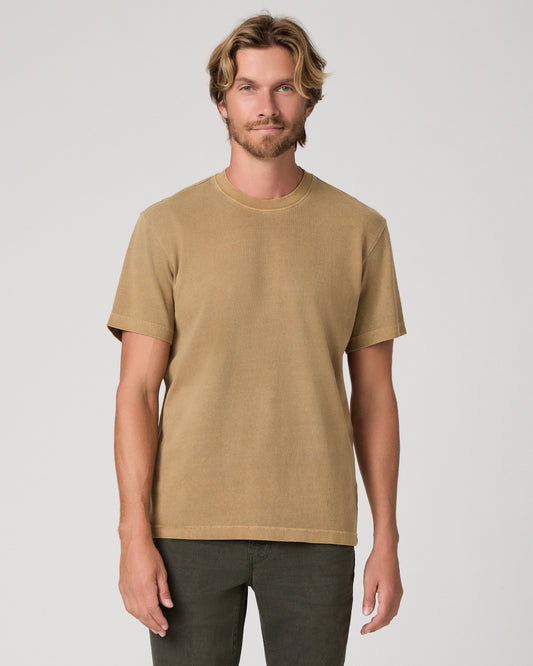 A man modeling a brown tee and dark green trousers against a light gray studio background.