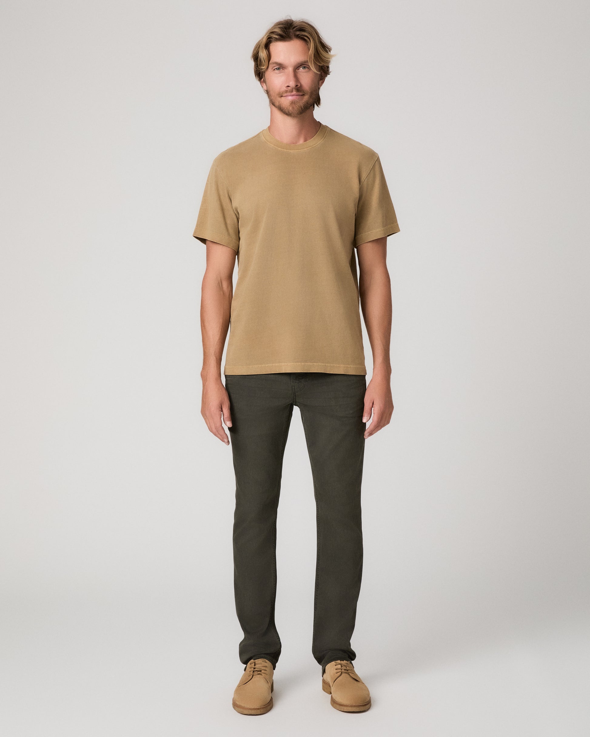 A man modeling a brown tee and dark green trousers paired with brown boots against a light gray studio background.