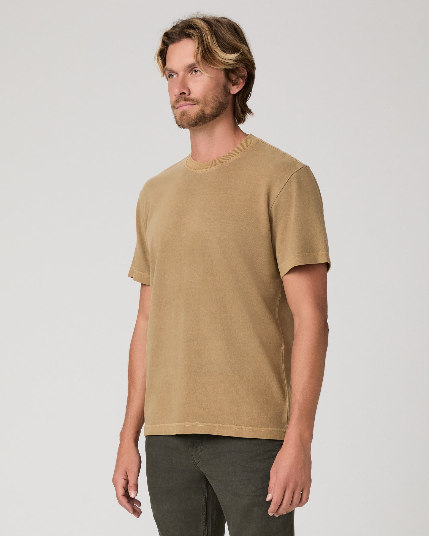 A man modeling a brown tee and dark green trousers against a light gray studio background.