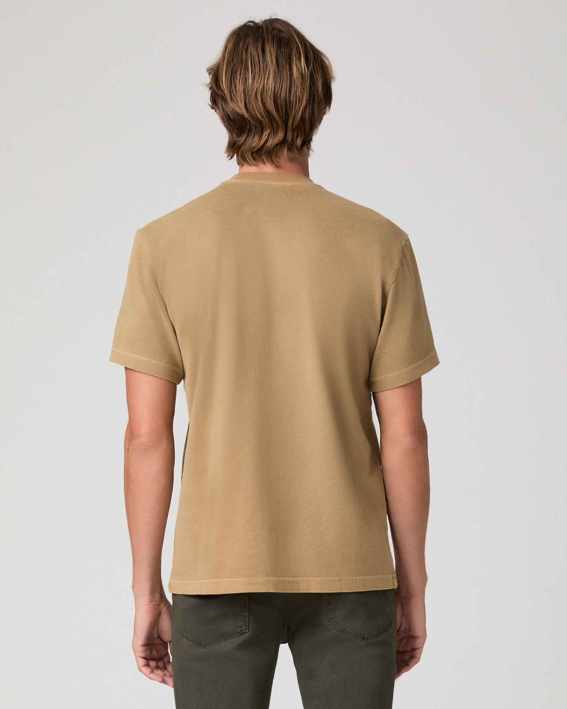 A man modeling a brown tee and dark green trousers against a light gray studio background.