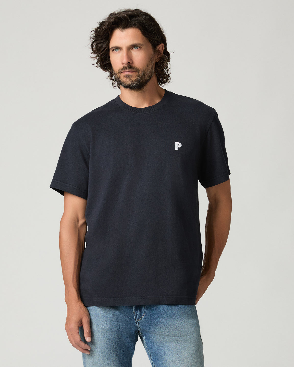 Cropped front view of a man with curly dark hair wearing a deep navy boxy-fit tee with a small white embroidered logo patch on the left chest, paired with light-wash jeans.