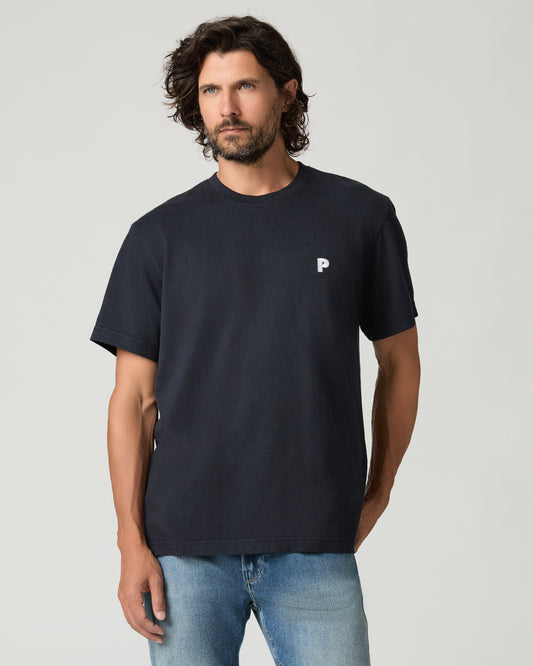 Cropped front view of a man with curly dark hair wearing a deep navy boxy-fit tee with a small white embroidered logo patch on the left chest, paired with light-wash jeans.