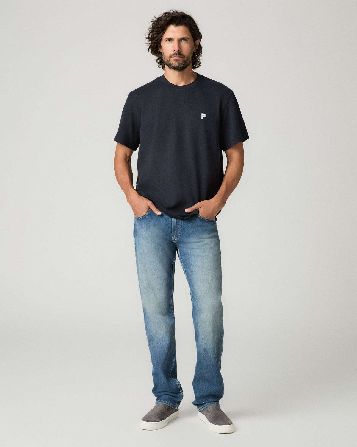 Full-length front view of a man walking with hands in his pockets, wearing a deep navy boxy tee with a small white chest logo, light-wash straight-leg jeans, and gray suede slip-on sneakers.
