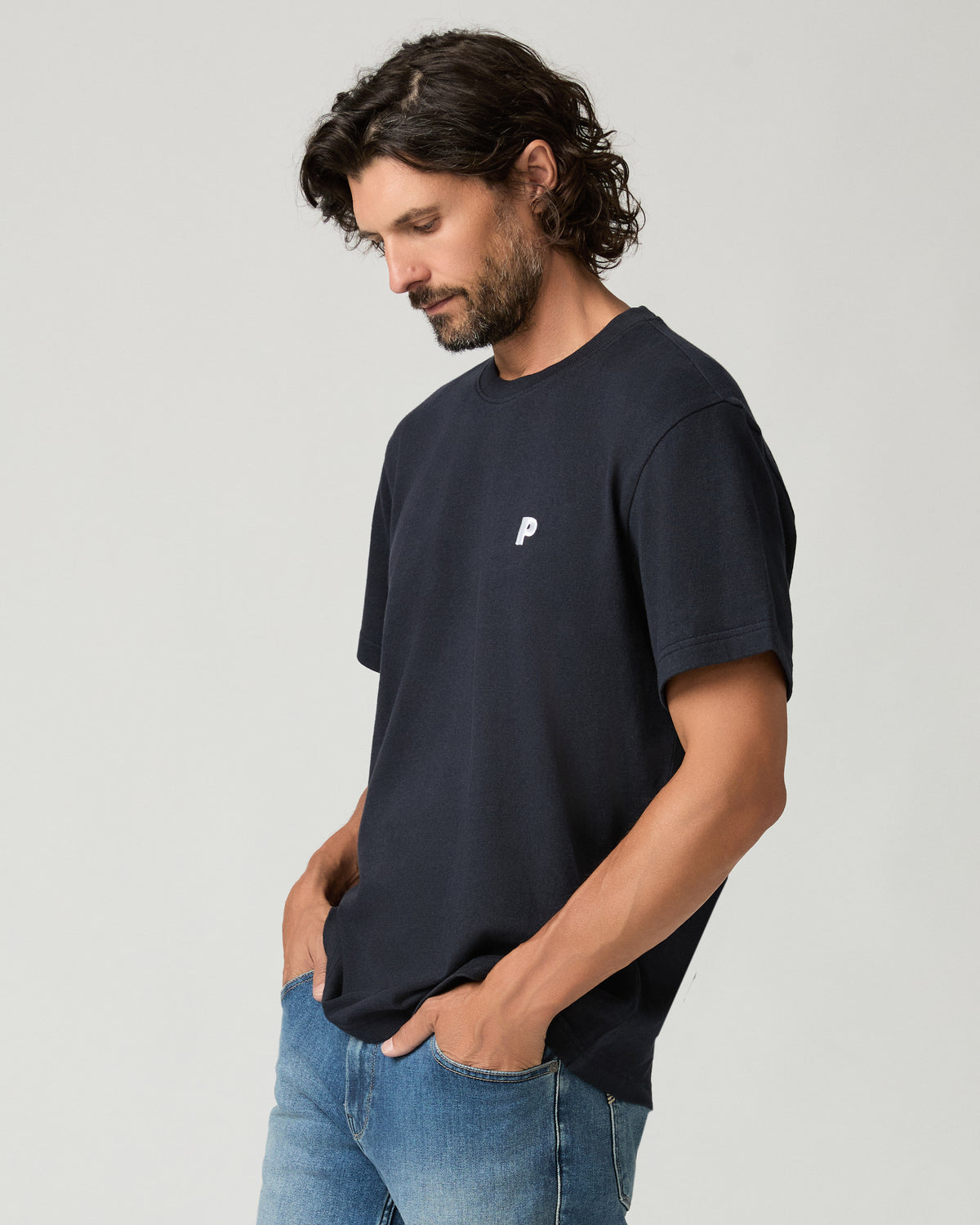 Three-quarter side view of a man looking down with one hand in his jeans pocket, wearing a deep navy boxy-fit tee with a small white chest logo and light-wash denim.
