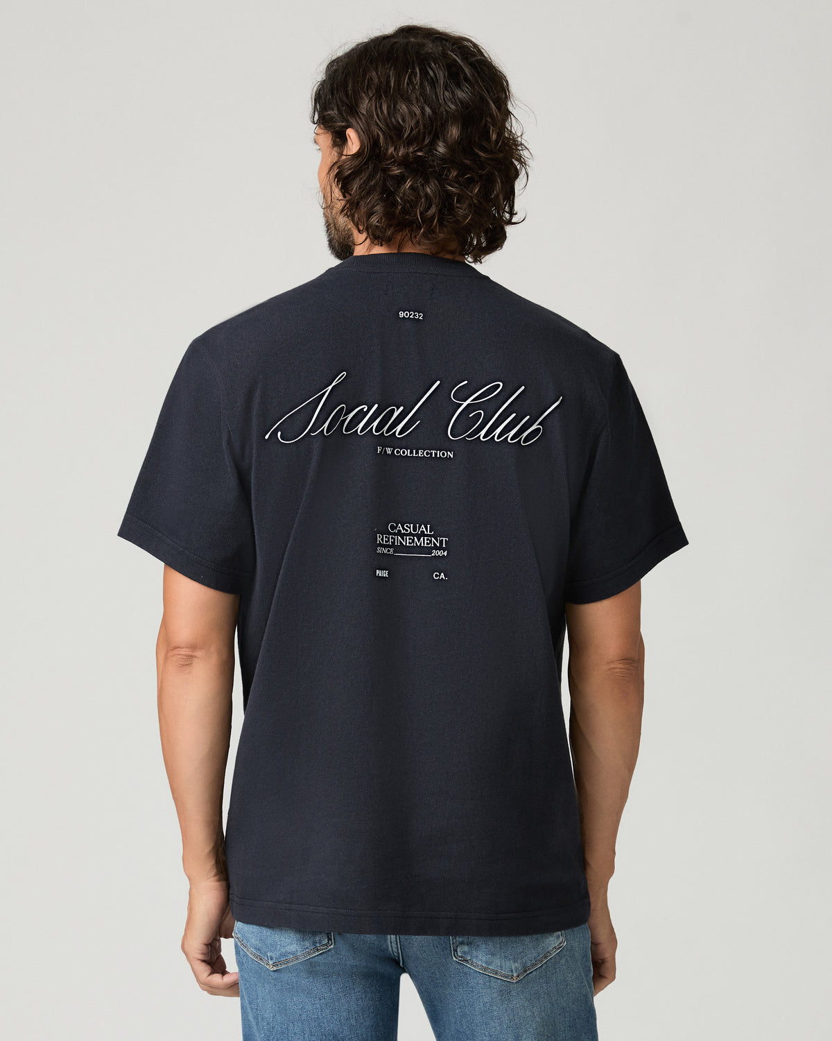 Back view of a man wearing a deep navy tee featuring a large 'Social Club' script graphic print across the upper back with smaller text beneath it.