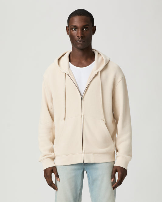 Man wearing a beige hoodie and light blue jeans on a white background