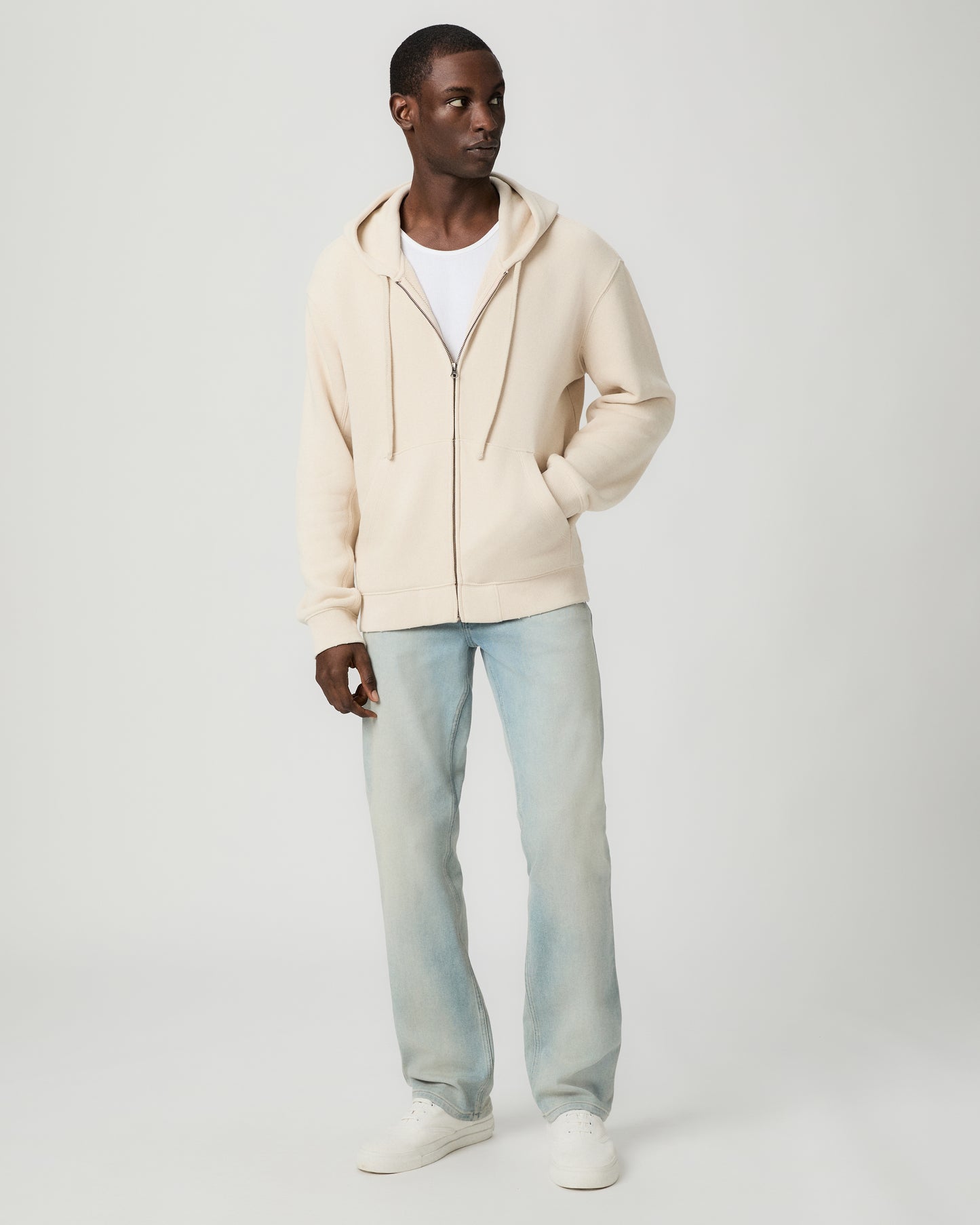 Man wearing a beige hoodie and light blue jeans on a white background