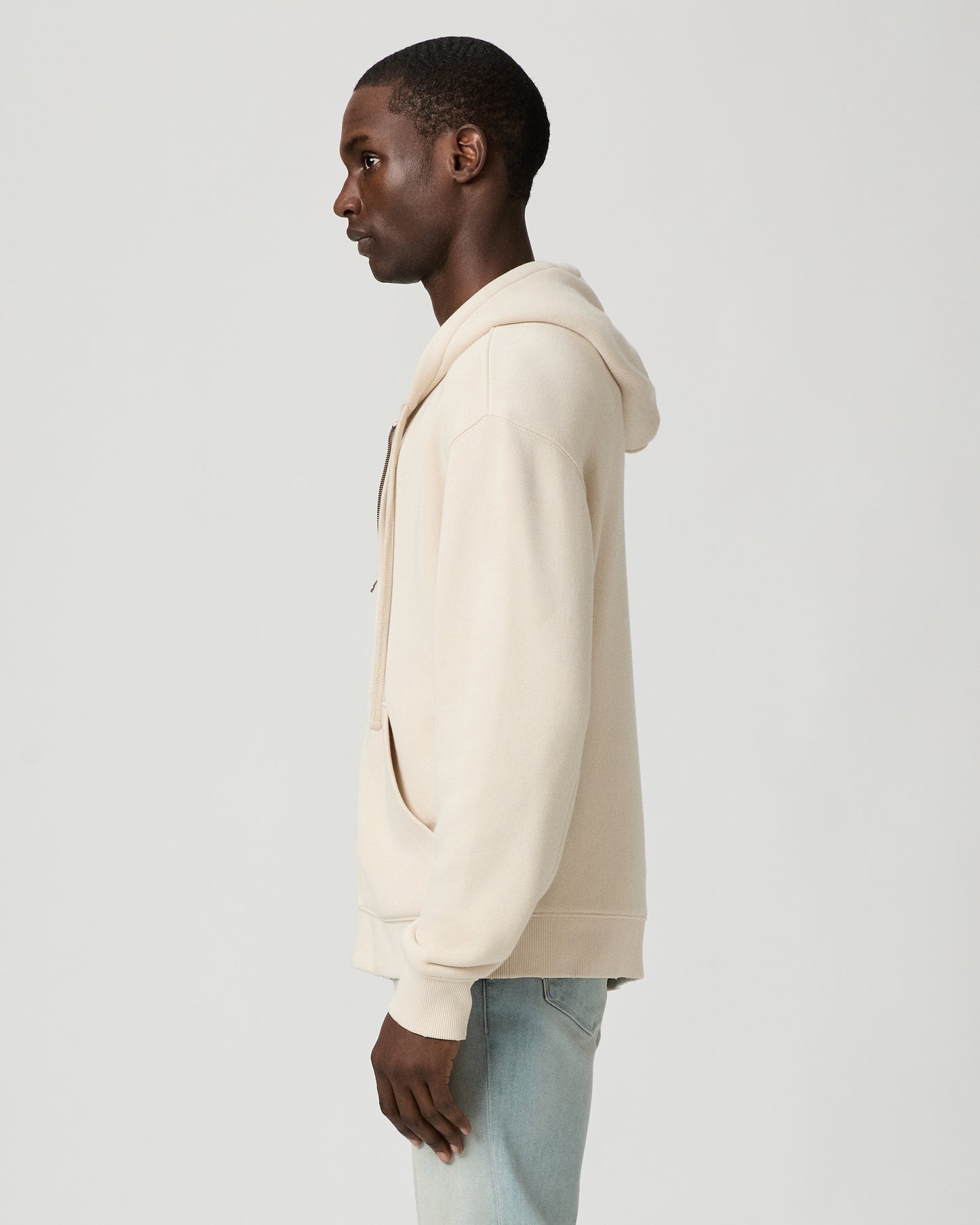 Man wearing a beige hoodie and light blue jeans on a white background