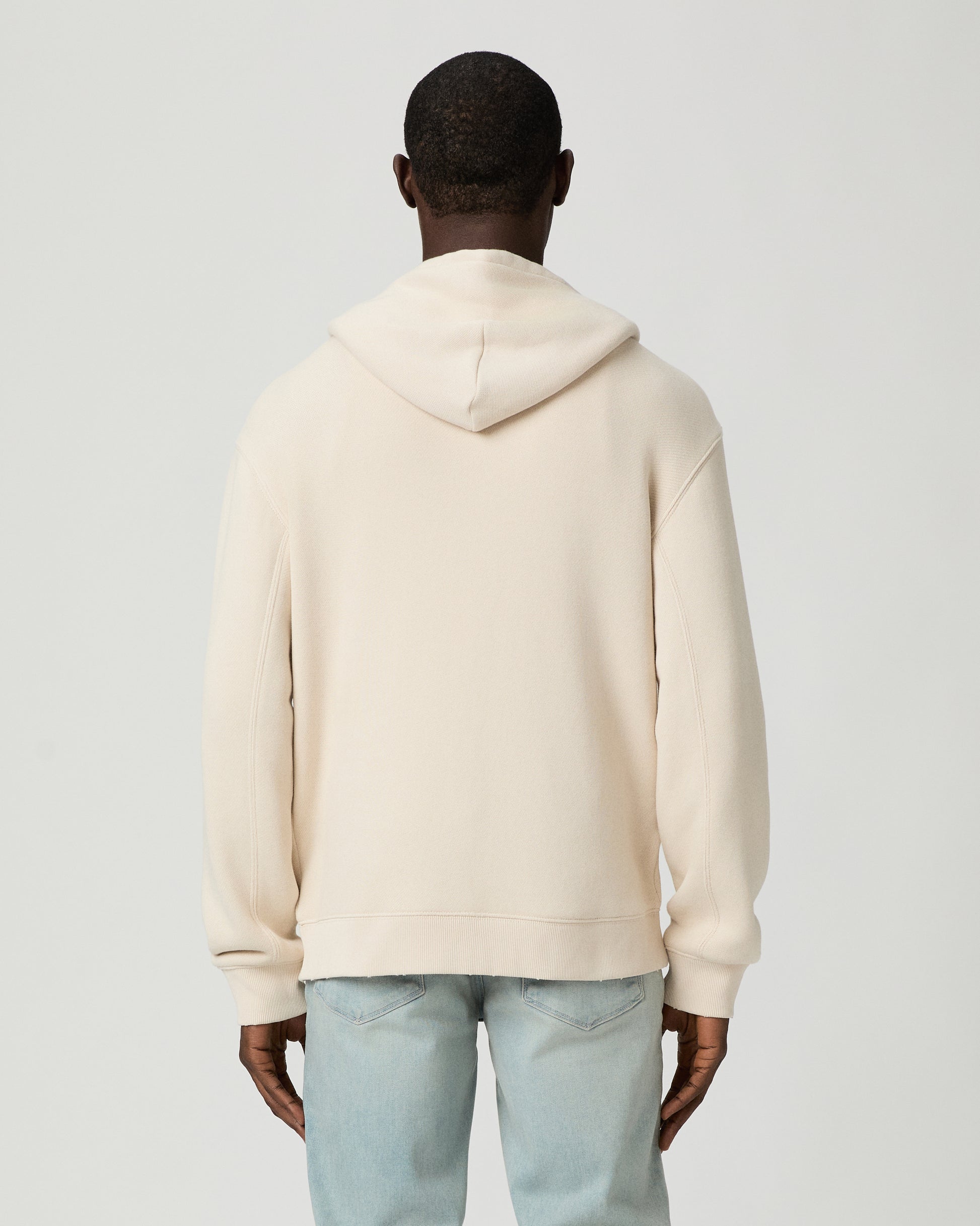 Man wearing a beige hoodie and light blue jeans on a white background
