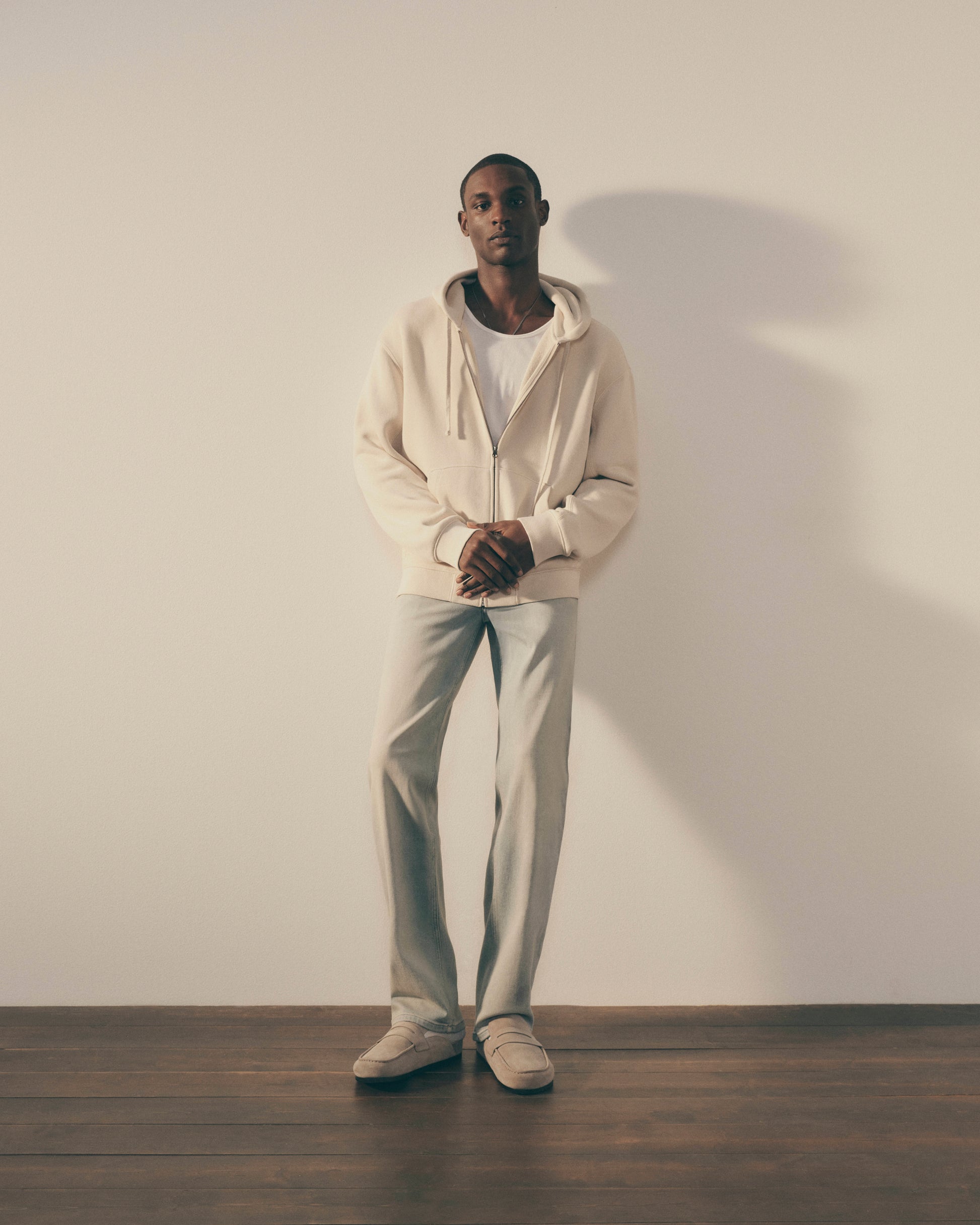 Man wearing a beige hoodie and pants standing against a white wall.