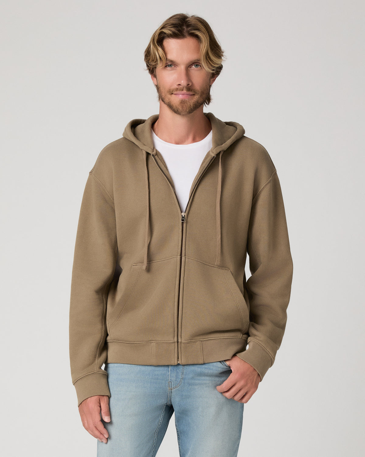 A man wearing a taupe-brown zip-up hoodie over a white t-shirt and light blue jeans, standing against a light gray studio background.