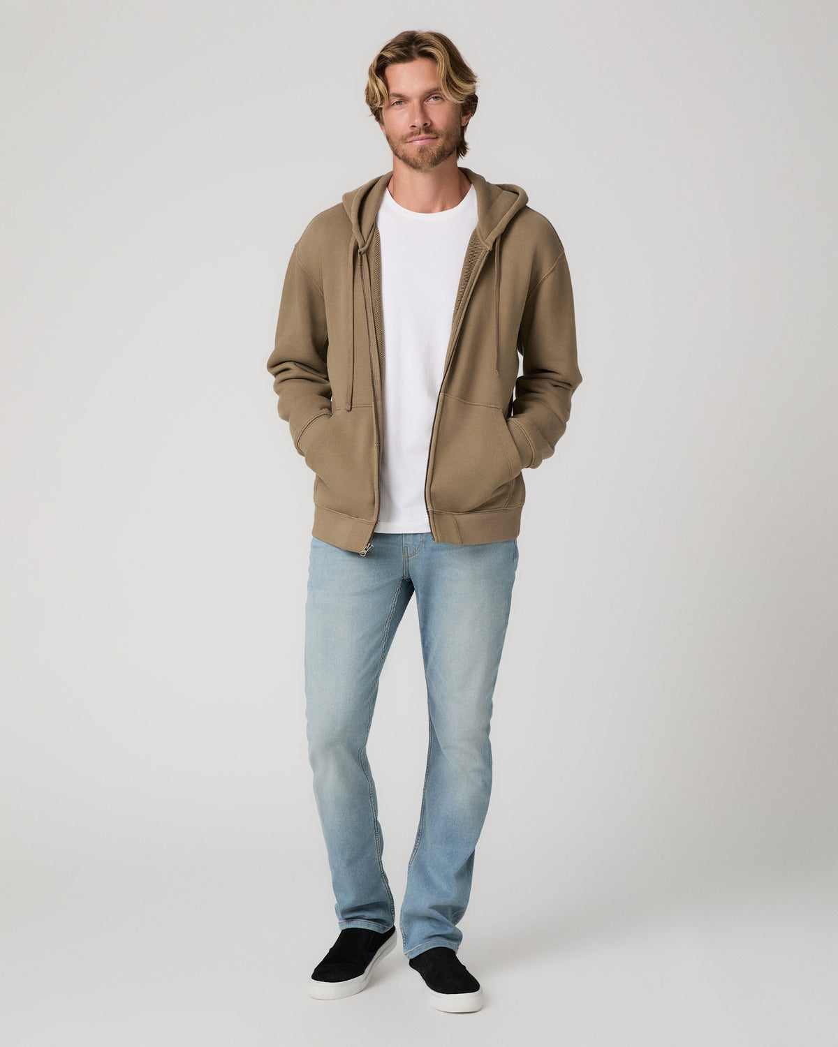 A man modeling a light brown zip-up hoodie over a white t-shirt, paired with light blue jeans and black slip-on shoes, against a plain light gray background.