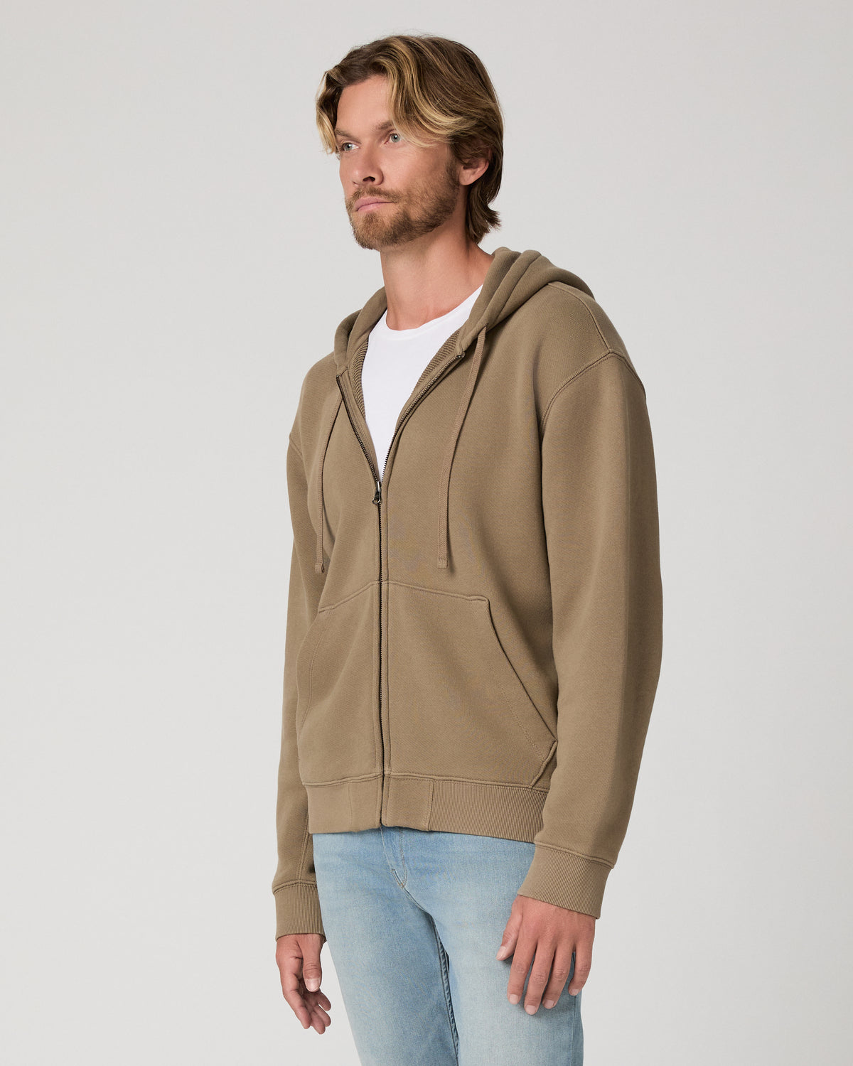 A man wearing a taupe-brown zip-up hoodie over a white t-shirt and light blue jeans, standing against a light gray studio background.
