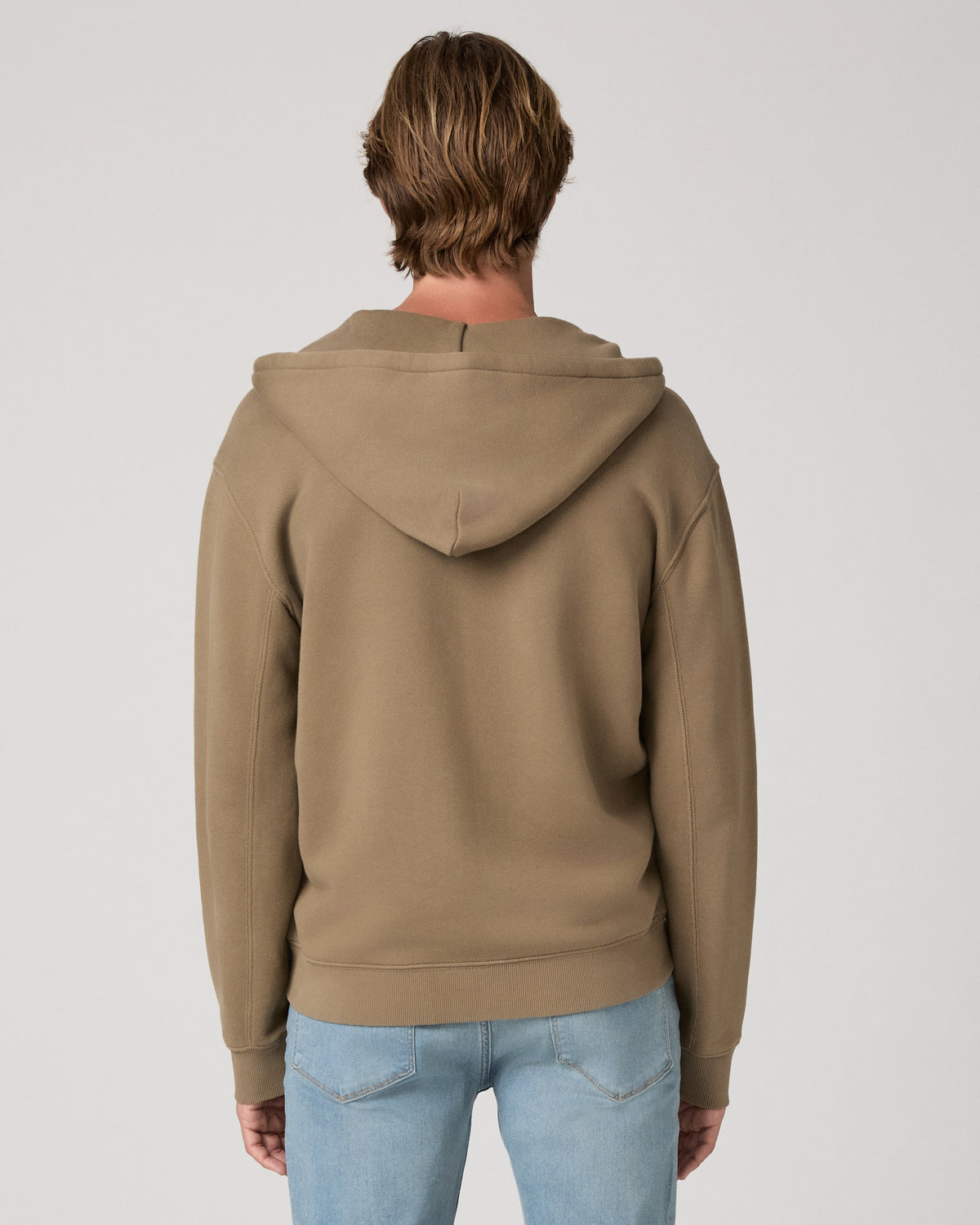 A man viewed from behind wearing a taupe brown pullover hoodie and light blue jeans, standing against a neutral light gray studio background.