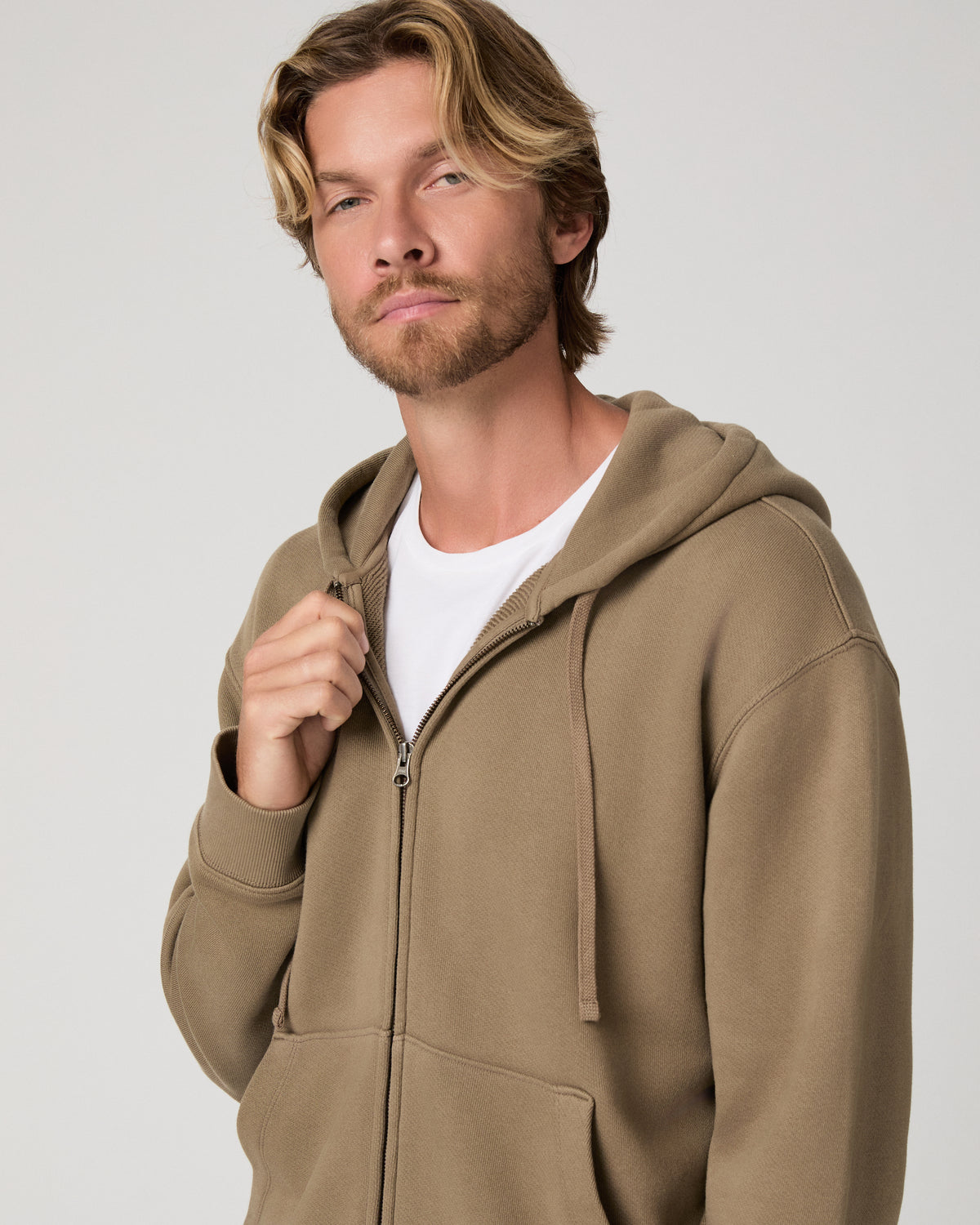 A man wearing a taupe brown zip-up hoodie over a white t-shirt, photographed against a neutral light gray studio background.