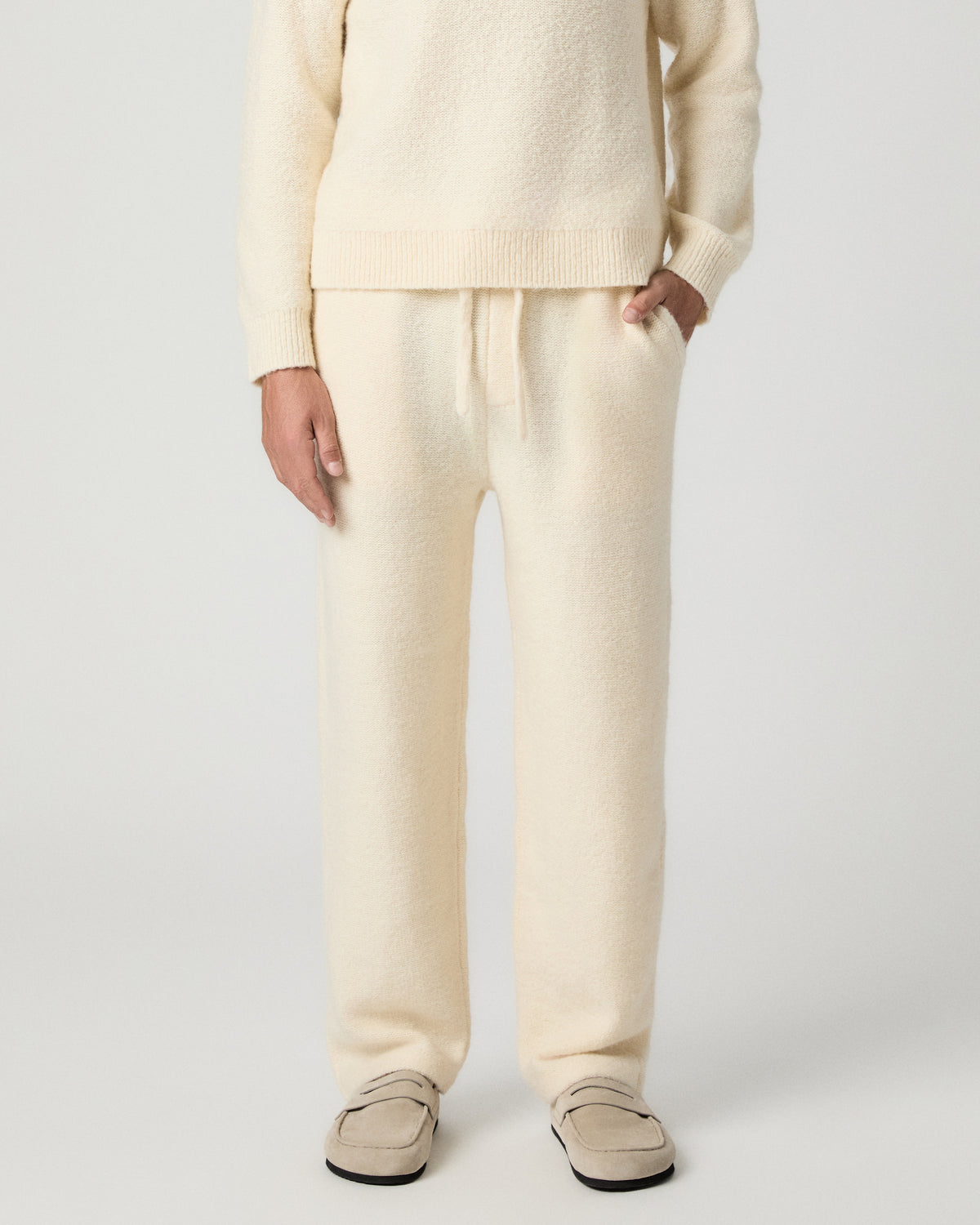 Front view from the waist down of the Willits Sweater Pant in Marfil, a cream knit drawstring pant, with one hand in the pocket, worn with a matching sweater and tan suede shoes