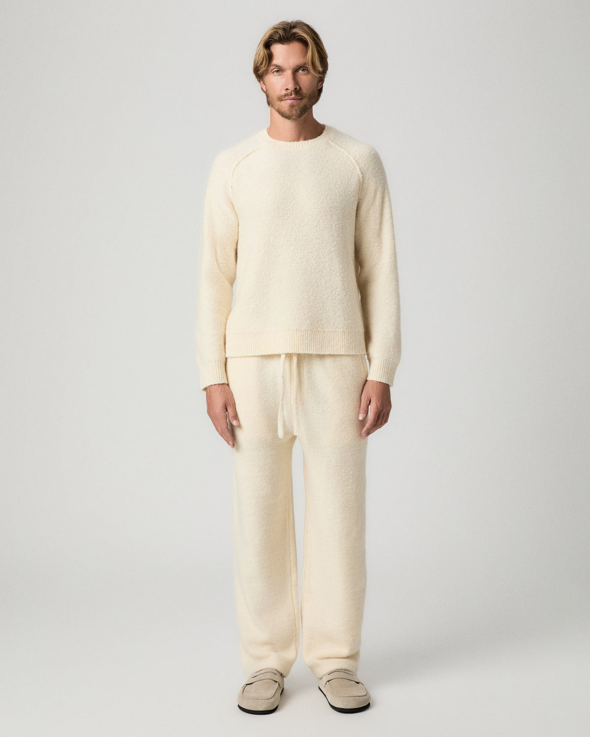 Full-length view of model standing with arms at his sides, wearing the Willits Sweater Pant in Marfil with a matching cream knit sweater and tan suede shoes
