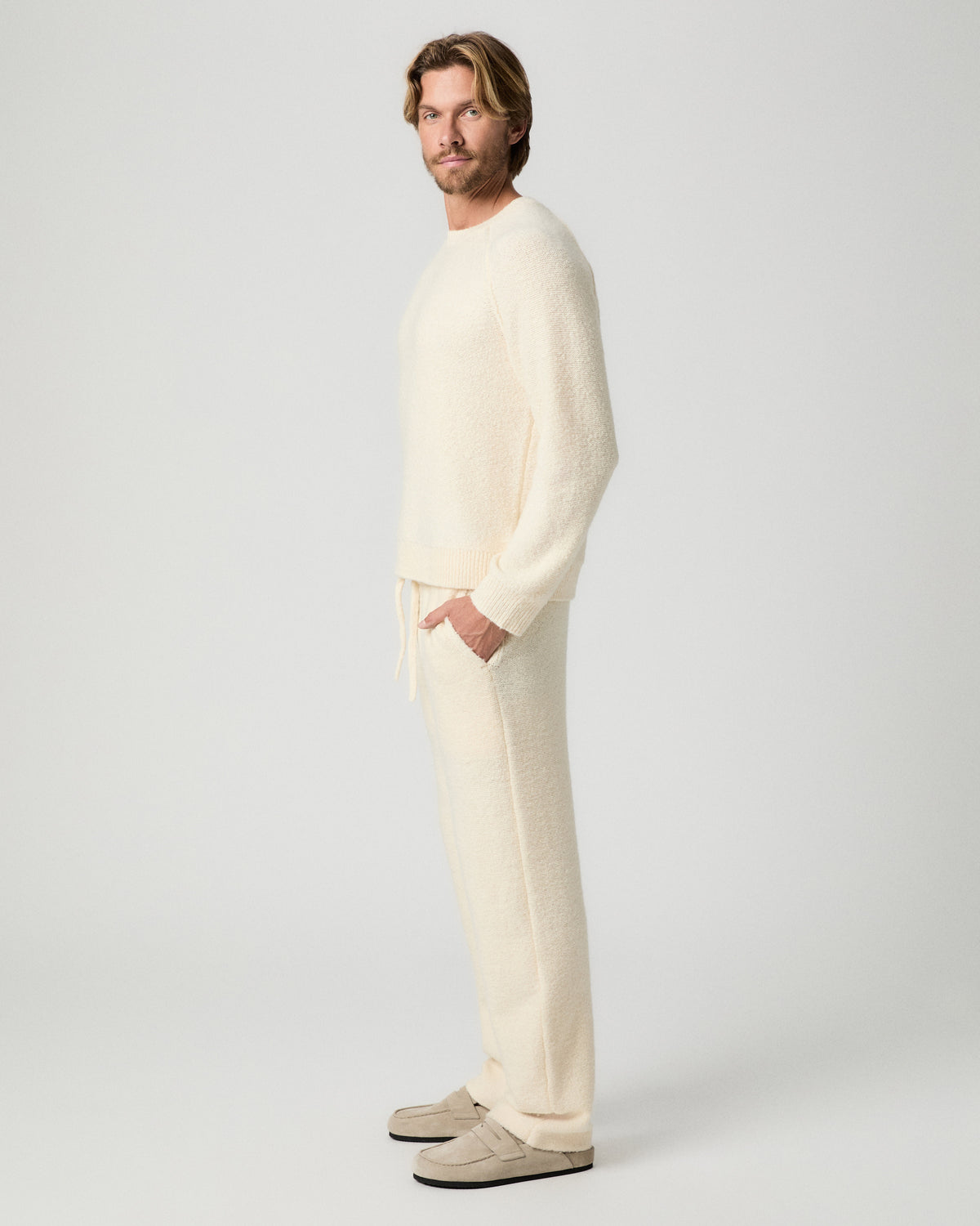 Side profile of model with one hand in his pocket, wearing the Willits Sweater Pant in Marfil with a matching cream knit sweater and tan suede shoes