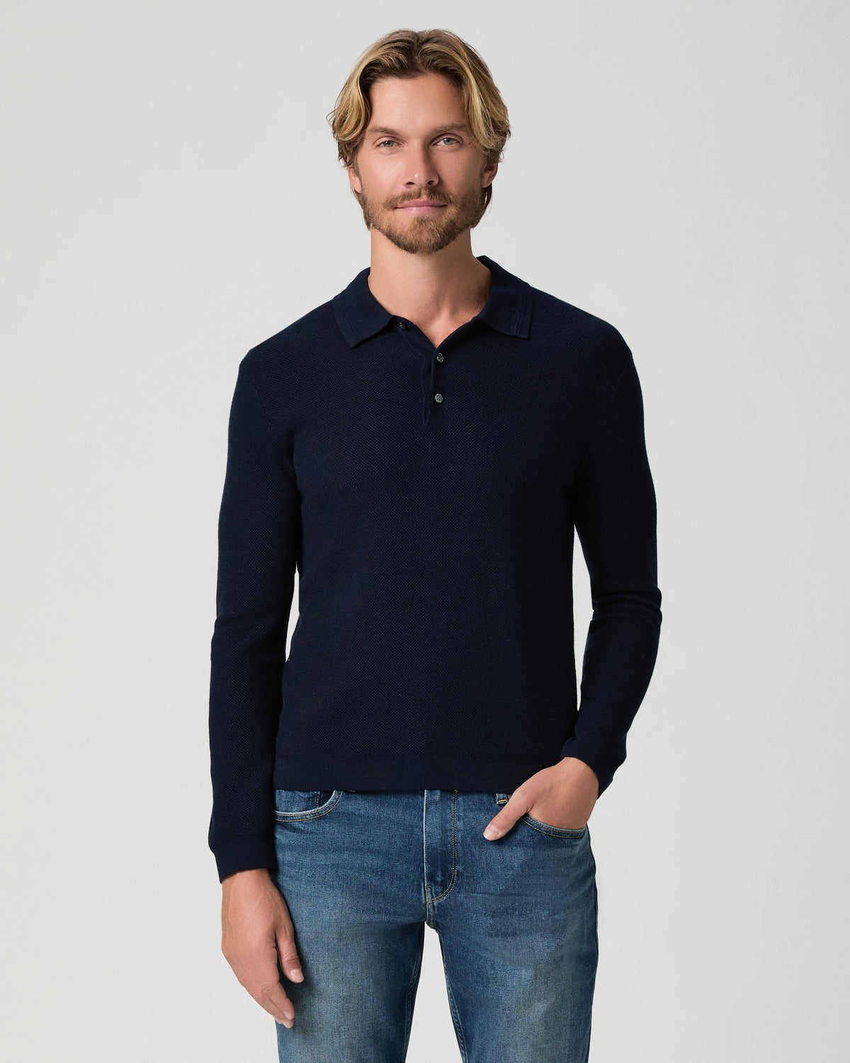 Model standing with one hand in his pocket, wearing the Selwyn Sweater Polo in Deep Anchor, a navy long-sleeve knit polo, with medium-wash jeans