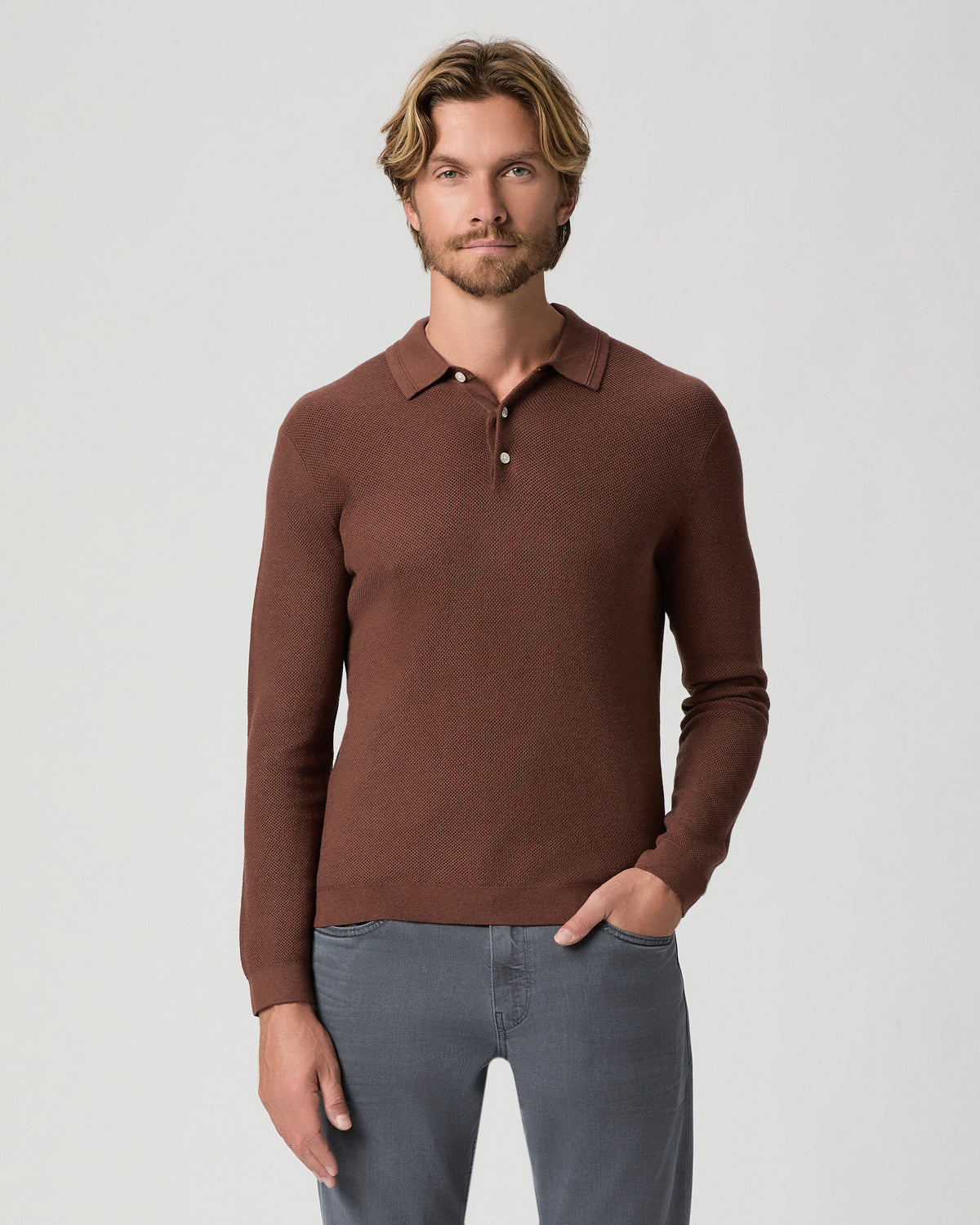 Model standing with one hand in his pocket, wearing the Selwyn Sweater Polo in Sorrel, a reddish-brown long-sleeve knit polo, with grey jeans