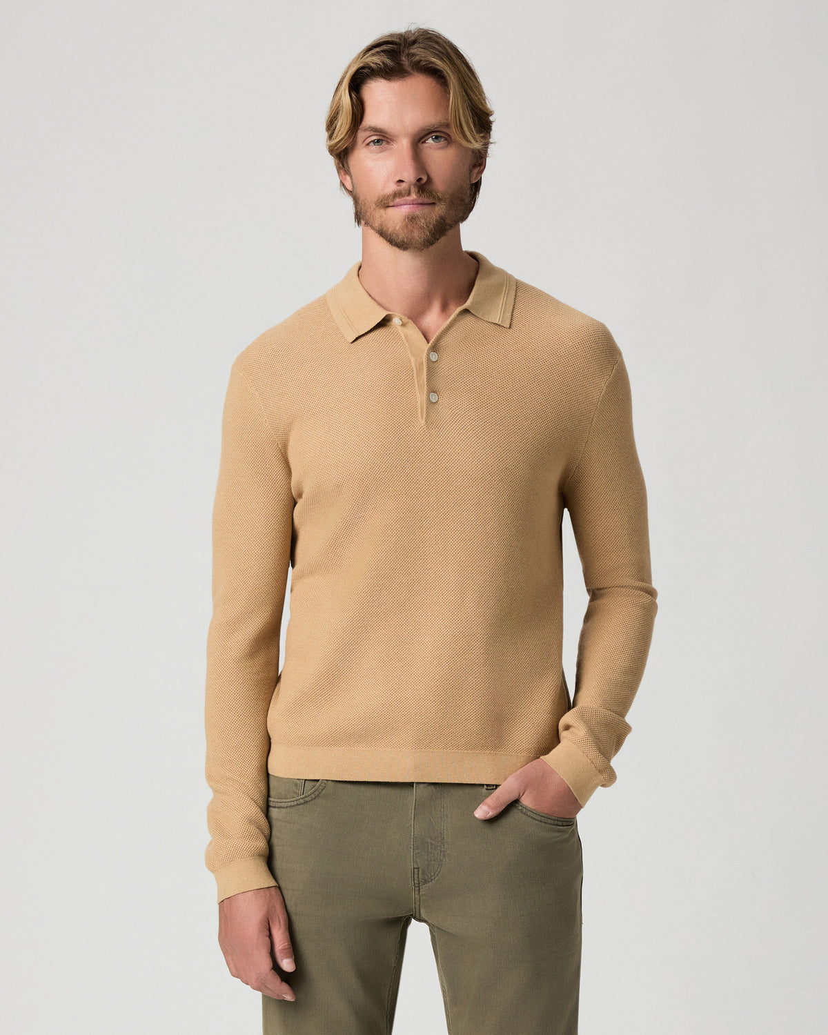 Model standing with one hand in his pocket, wearing the Selwyn Sweater Polo in Brown Russet, a tan long-sleeve knit polo, with olive pants