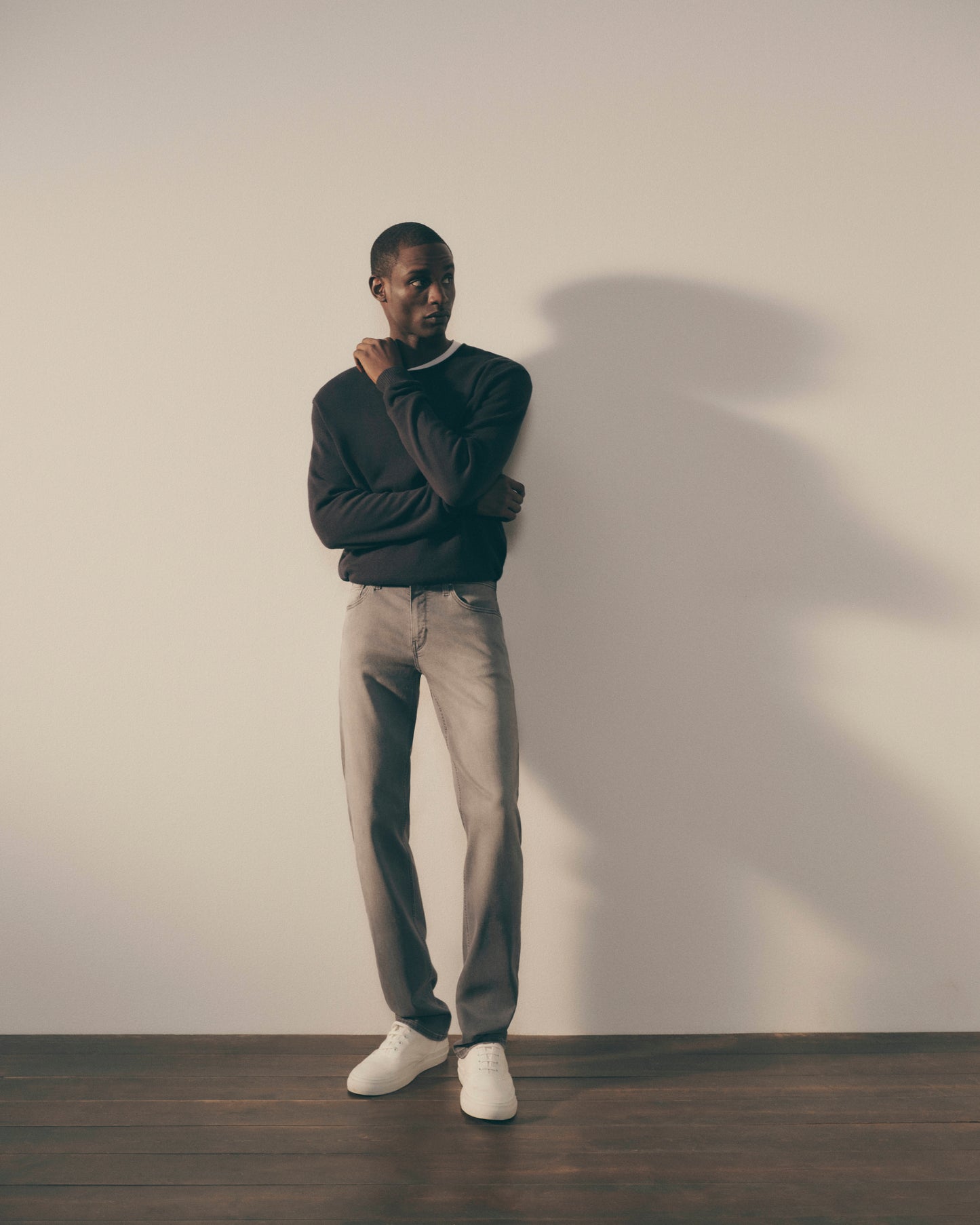 Man wearing a dark sweater, light jeans, and white shoes against a plain wall.