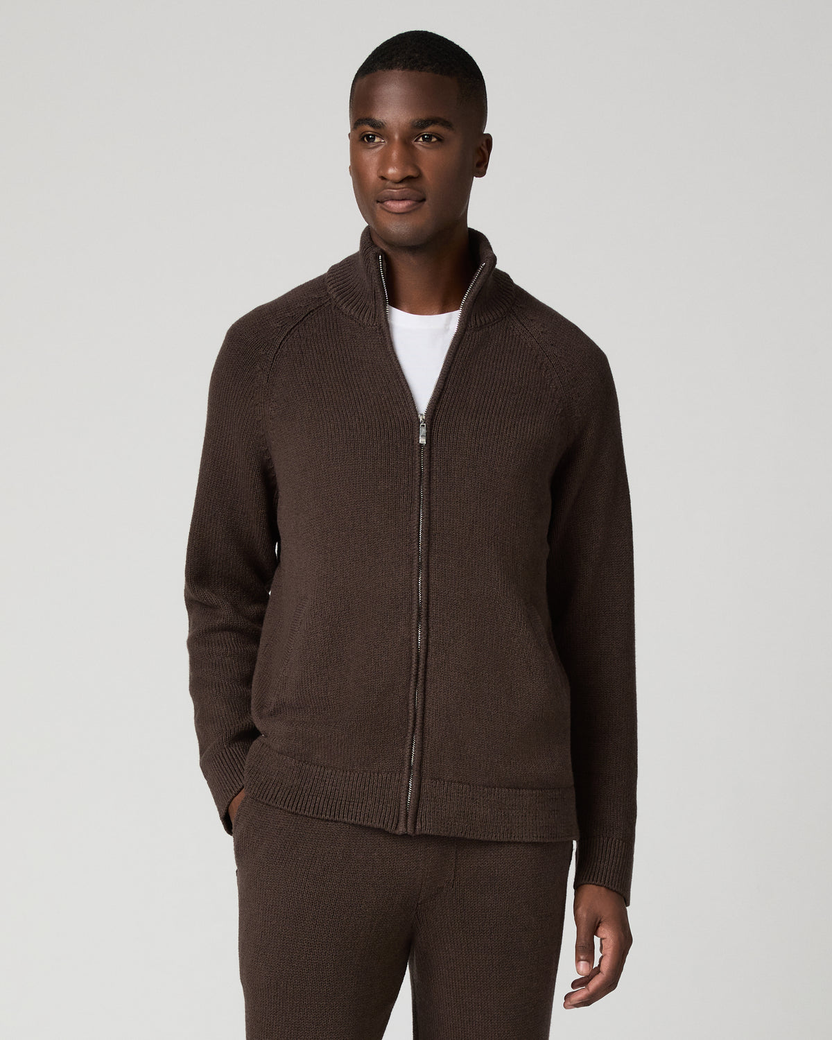 Full-length front view of a man wearing a chocolate brown zip-up cardigan with a stand collar over a white t-shirt, paired with matching brown sweatpants, arms relaxed at his sides.