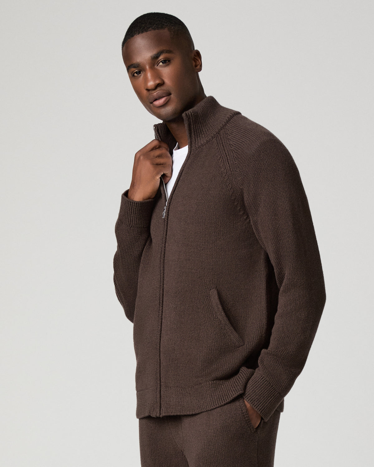 Three-quarter view of a man wearing the chocolate brown zip-up cardigan, one hand adjusting the ribbed stand collar and the other tucked in a front pocket.