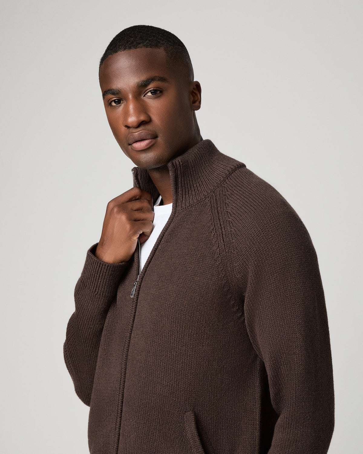 Close-up front view of a man wearing the chocolate brown zip-up cardigan, one hand adjusting the ribbed stand collar near the zipper pull, a white t-shirt visible underneath.