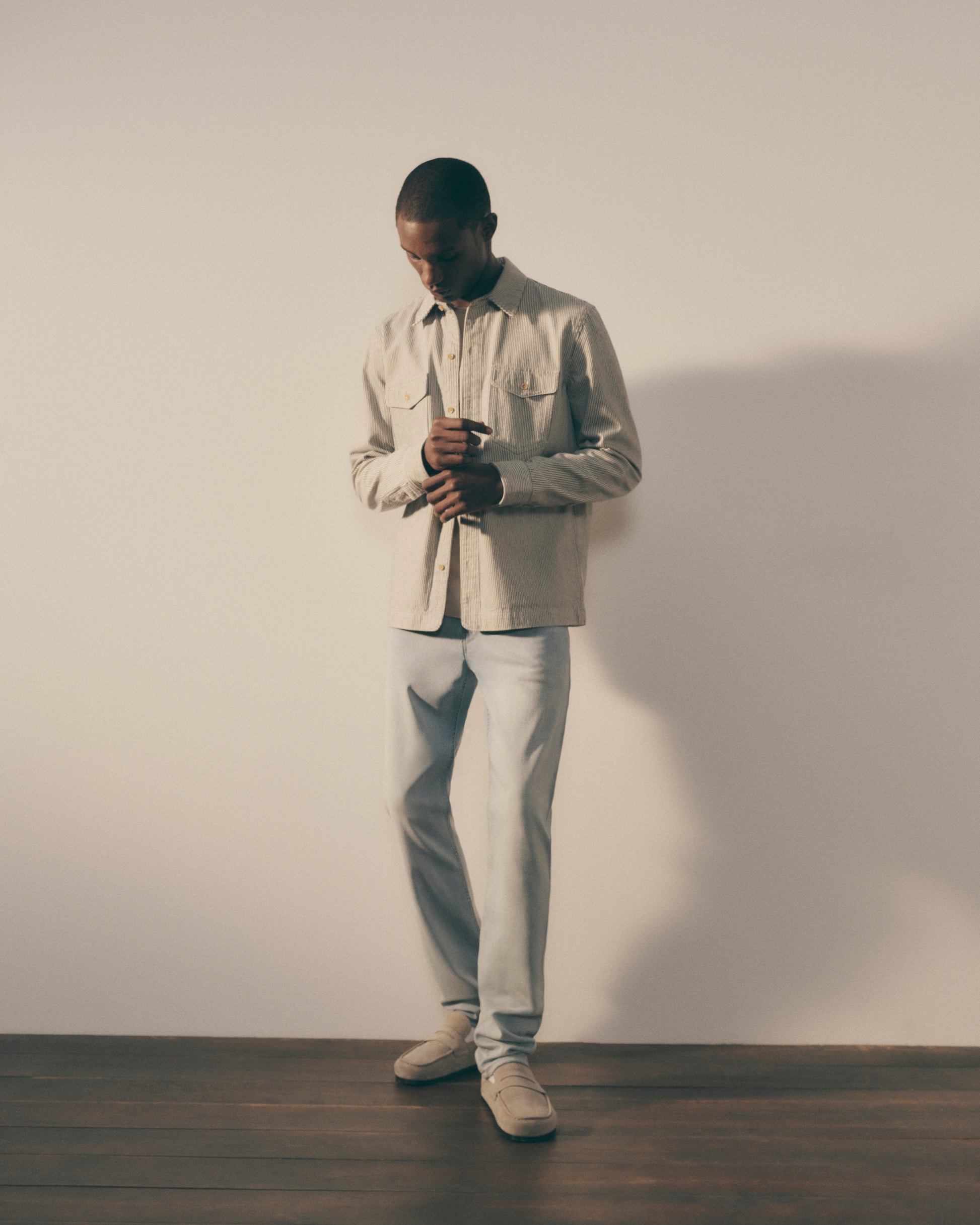 Man wearing a beige shirt and light gray pants against a plain wall.