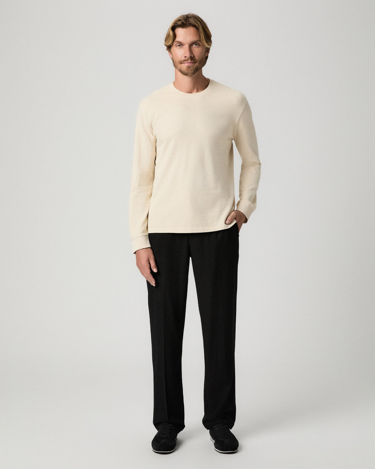 Full-length view of model with one hand in his pocket, wearing a cream long-sleeve sweater with the Tiano Pant in Black and black shoes