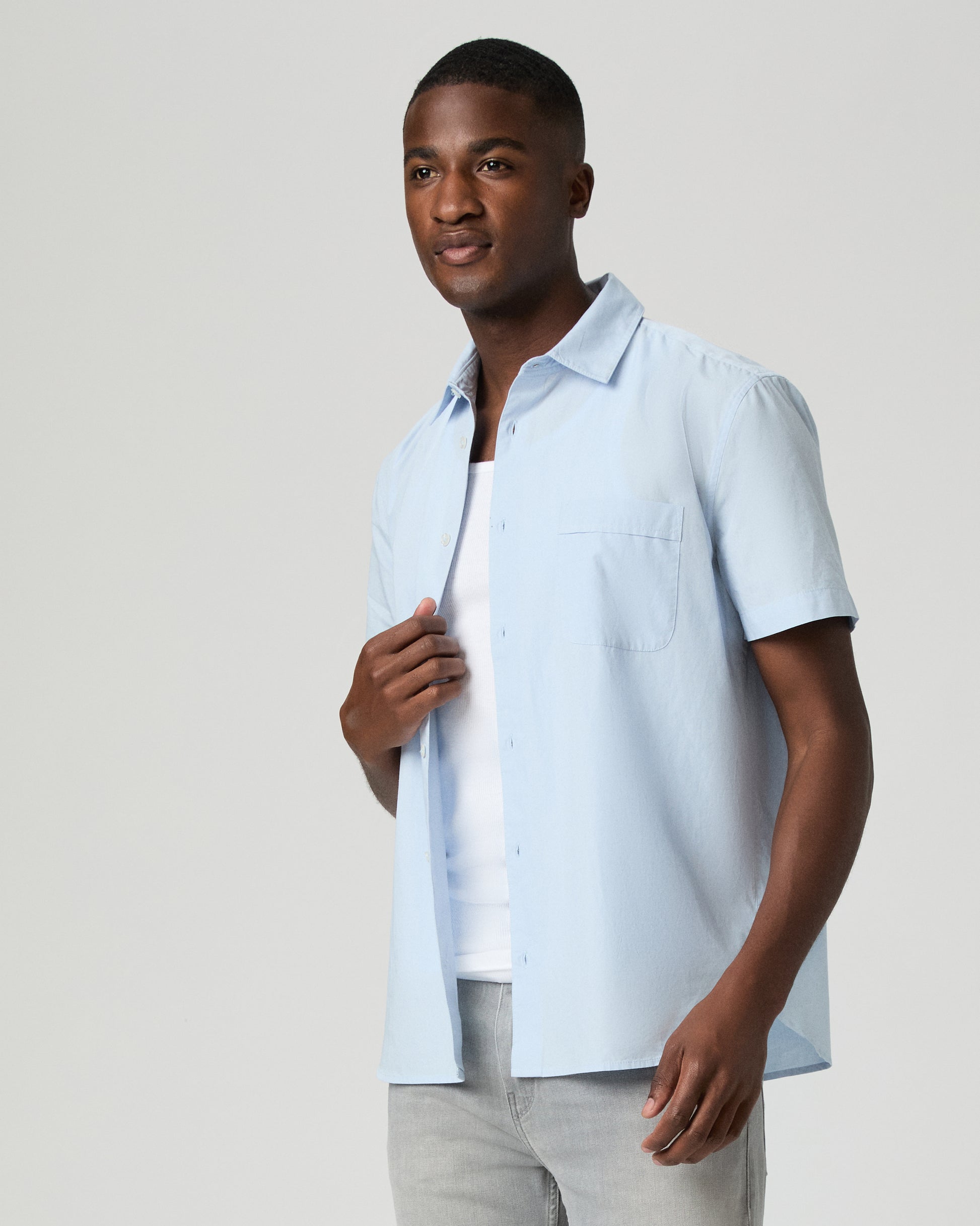Man wearing a light blue short-sleeve shirt over a white t-shirt on a plain background