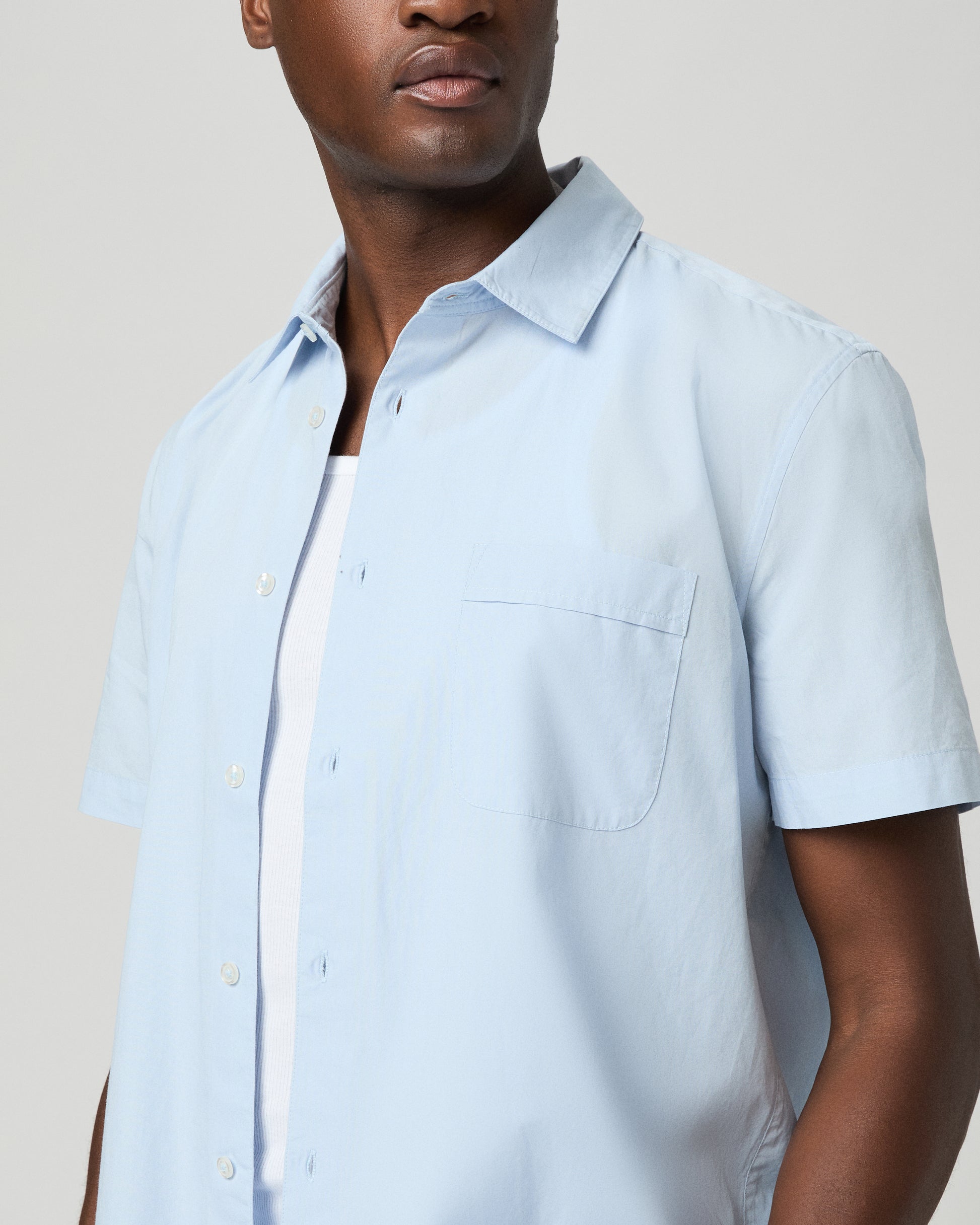 Man wearing a light blue short-sleeve shirt over a white t-shirt on a plain background