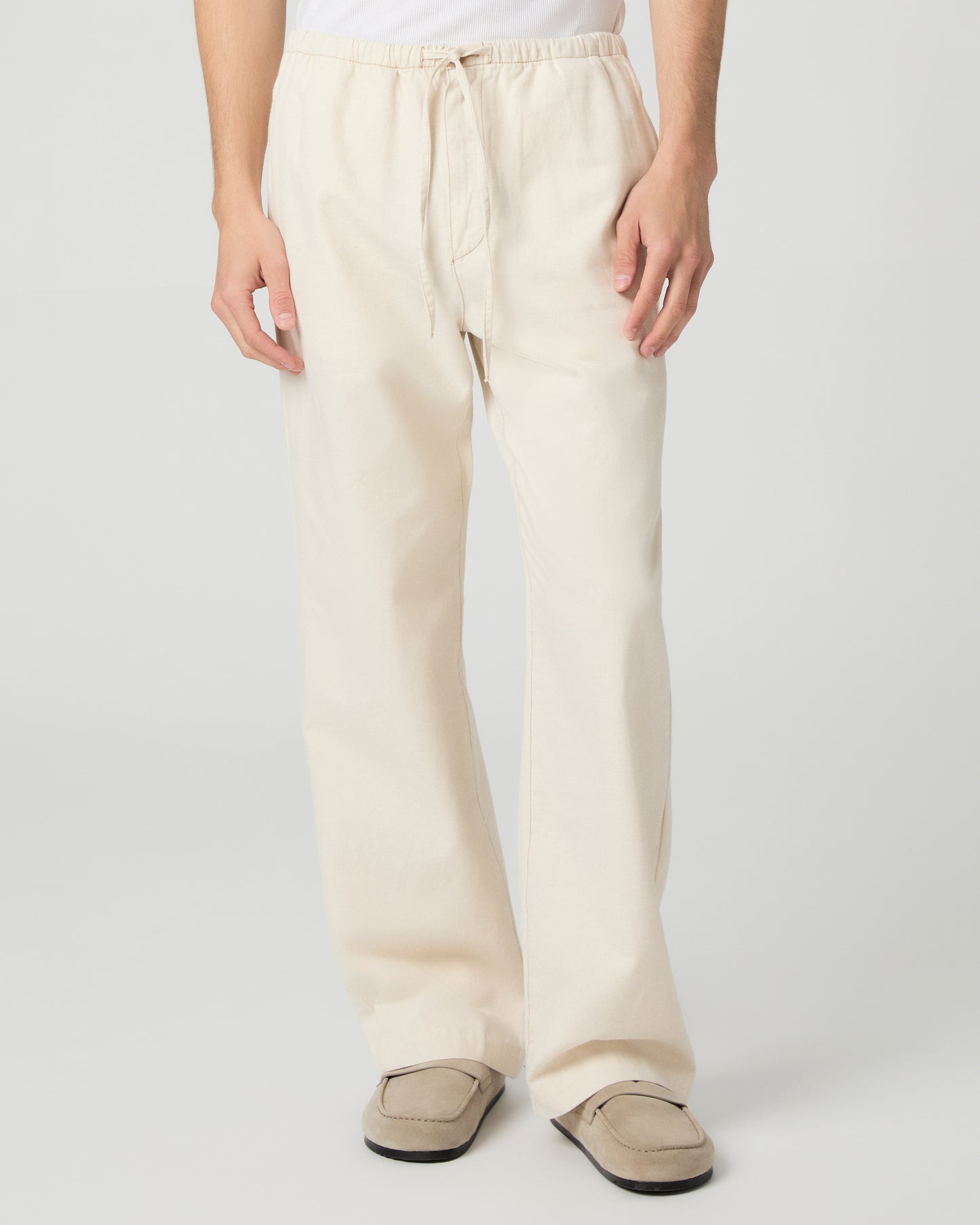 Man wearing a white tee and beige linen pants paired with grey shoes on a white background