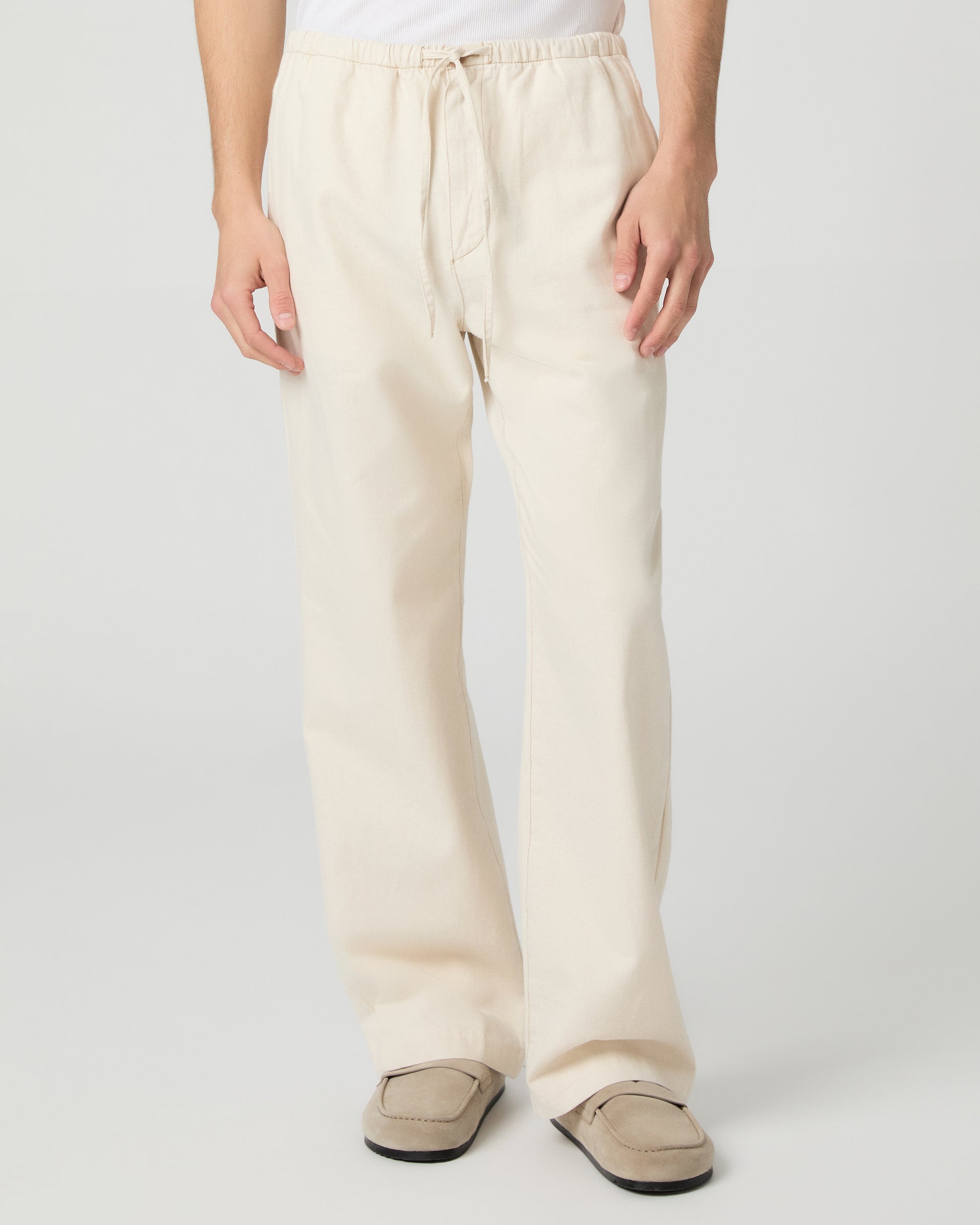 Man wearing a white tee and beige linen pants paired with grey shoes on a white background