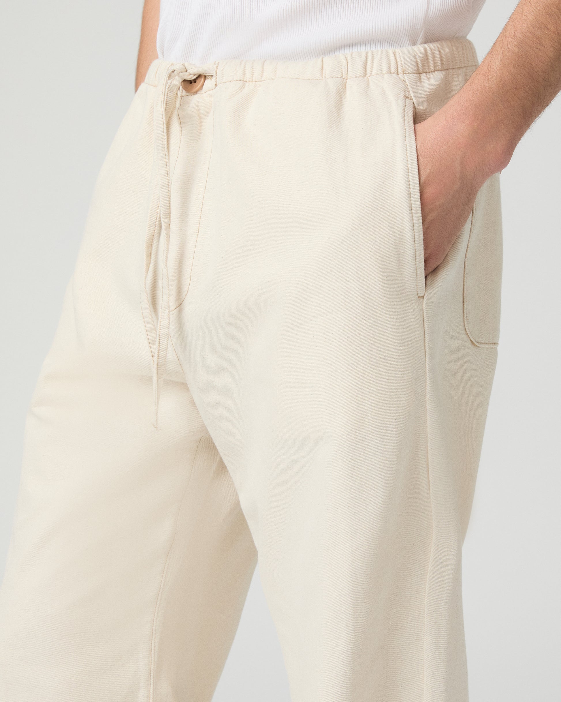 Man wearing a white tee and beige linen pants paired with grey shoes on a white background
