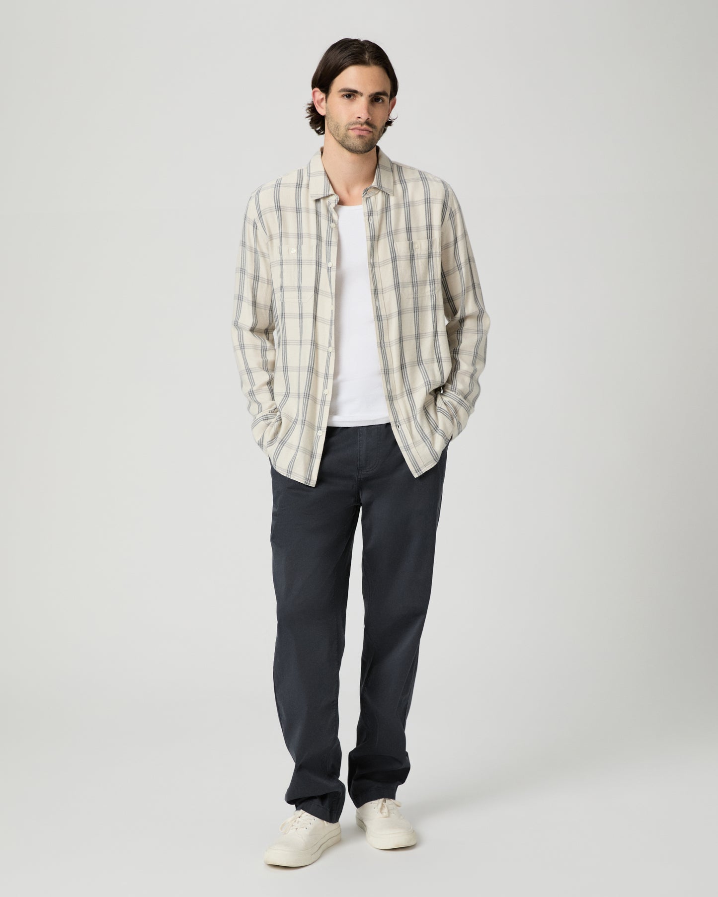 Man wearing a plaid shirt, white t-shirt, dark pants, and white shoes on a white background