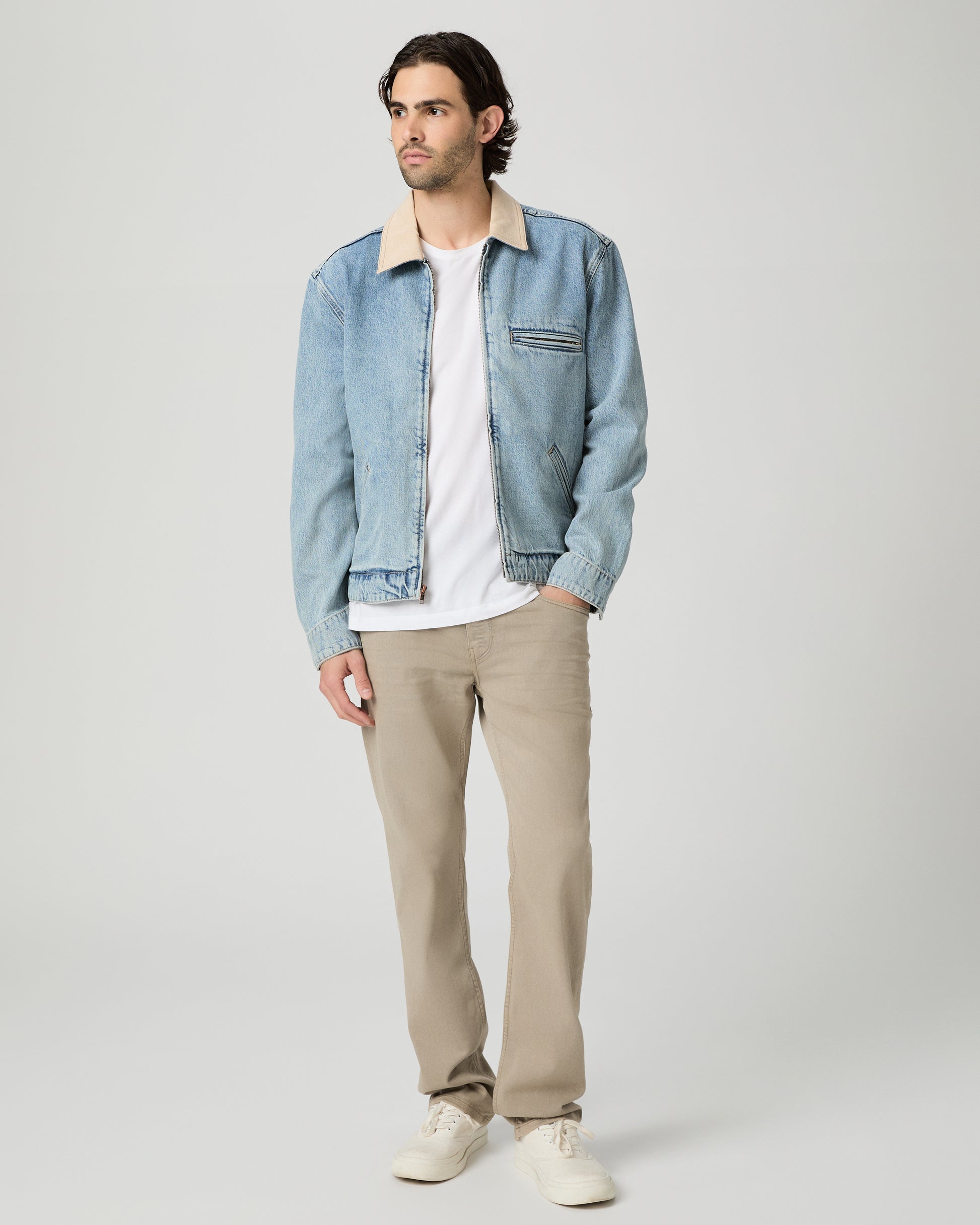 Man wearing a light blue denim jacket over a white shirt and beige pants on a gray background