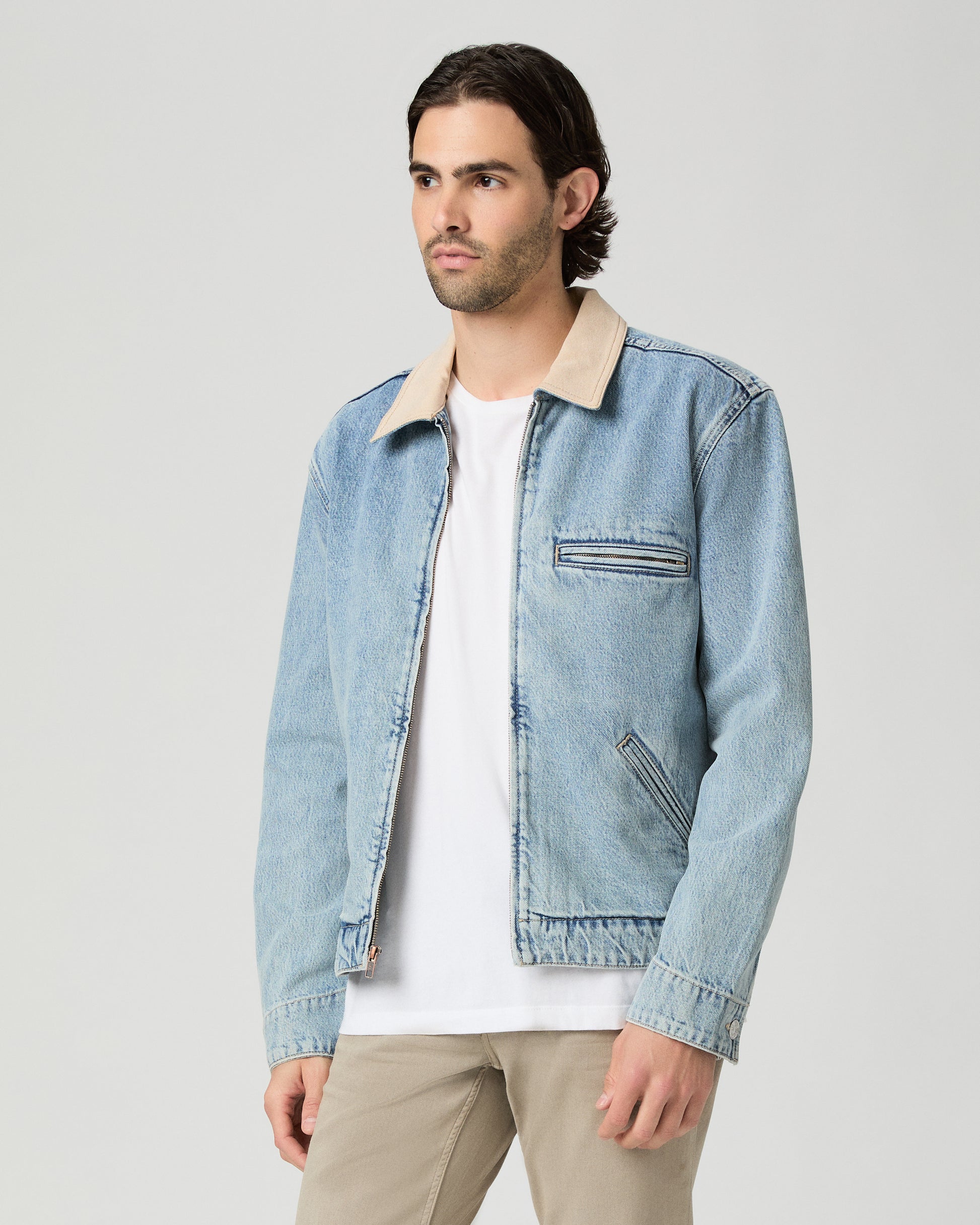 Man wearing a light blue denim jacket over a white shirt and beige pants on a gray background