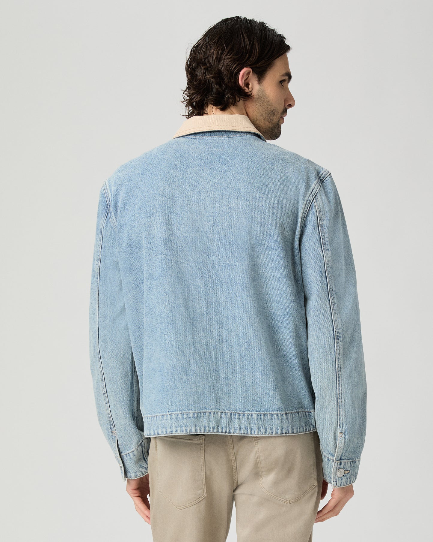 Man wearing a light blue denim jacket over a white shirt and beige pants on a gray background